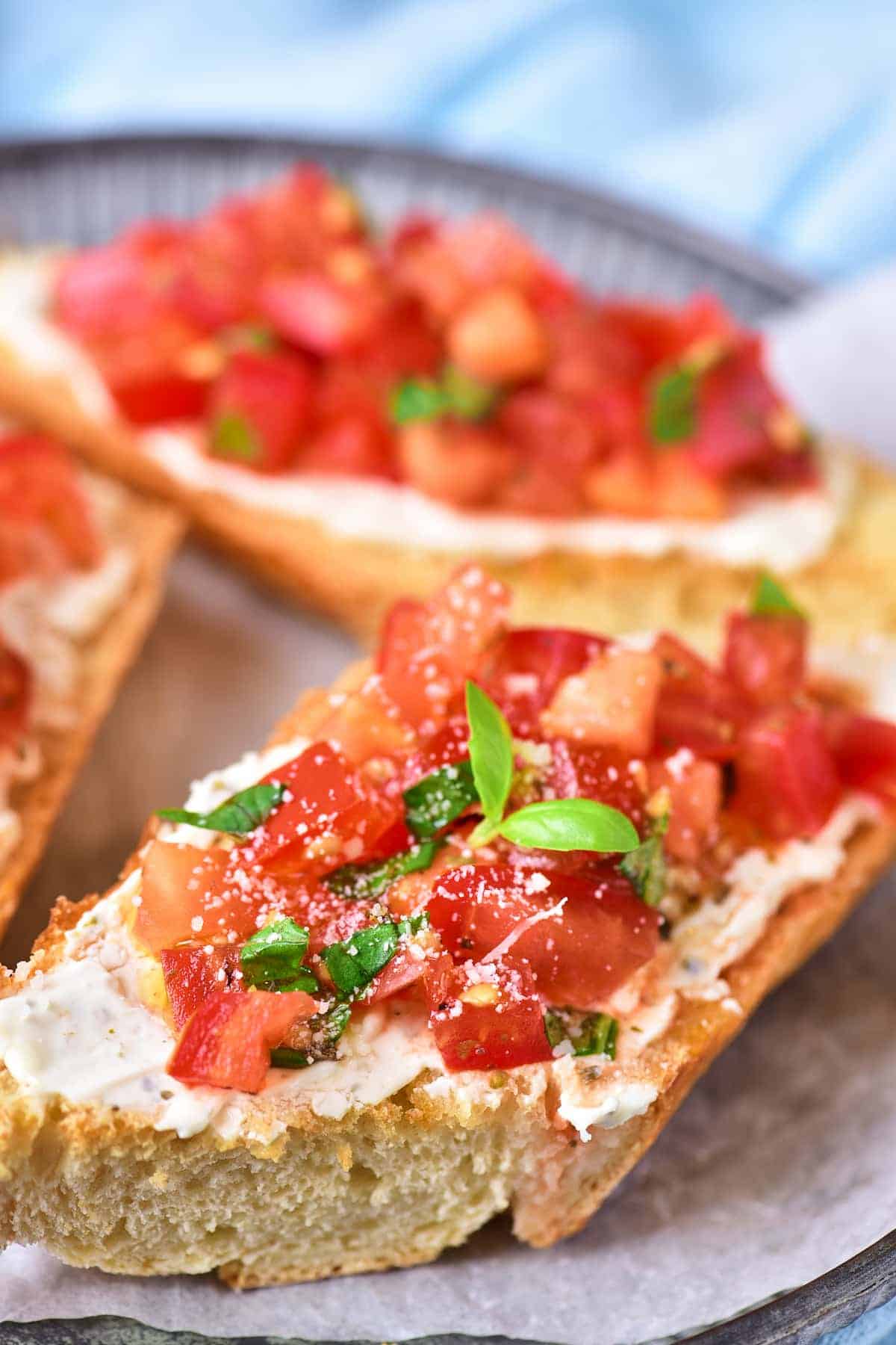 A close-up of bruschetta topped with diced tomatoes, herbs, grated cheese, and a layer of spread on sliced toasted bread.