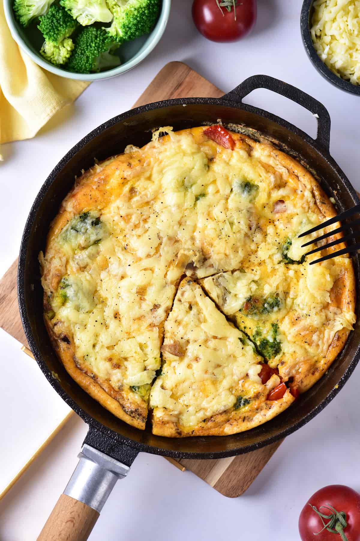 A sliced vegetable and cheese frittata in a black skillet, with a fork lifting one piece. Surrounding are fresh broccoli, tomatoes, and a bowl of grated cheese.