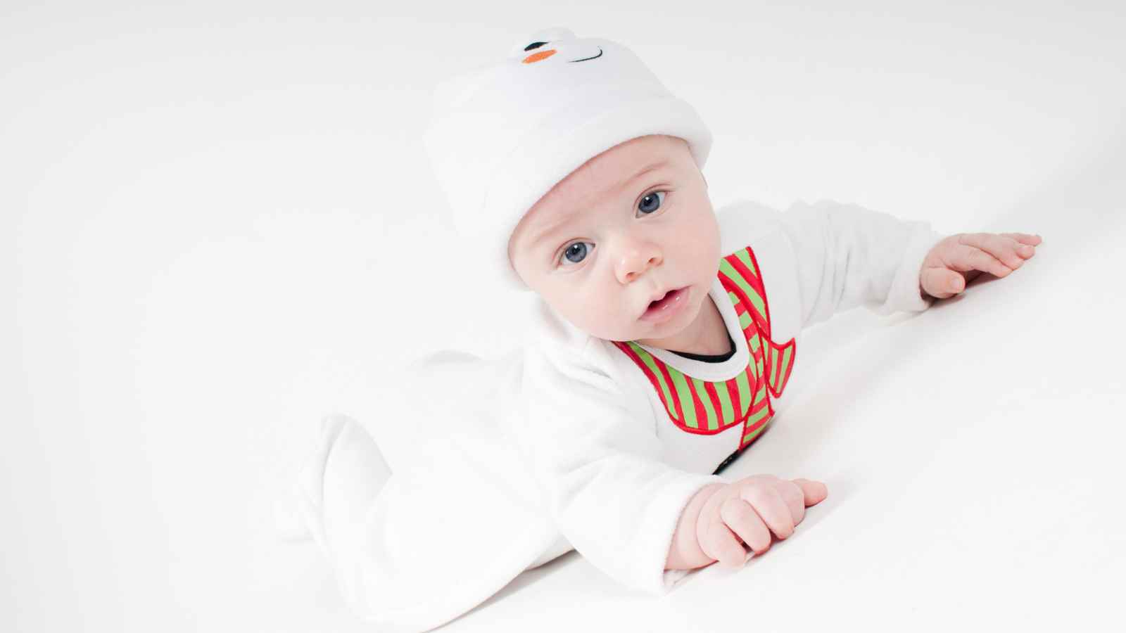 Baby lying on stomach, wearing a white outfit and hat with a snowman face, looking toward the camera against a white background.