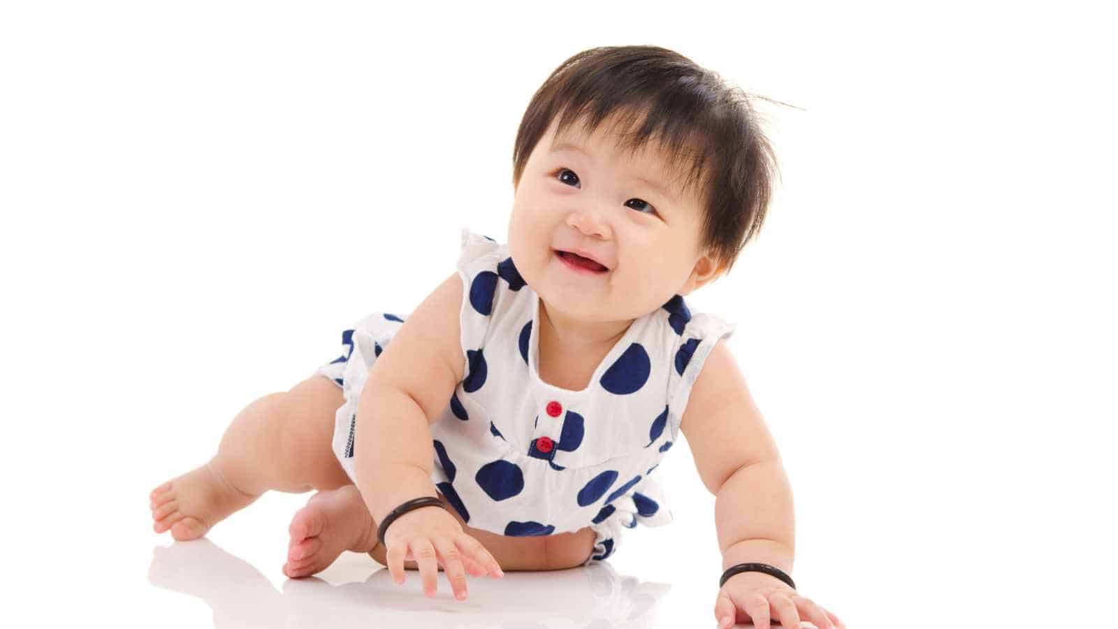 A baby wearing a white outfit with blue polka dots is crawling on a white surface, smiling and looking slightly upward.