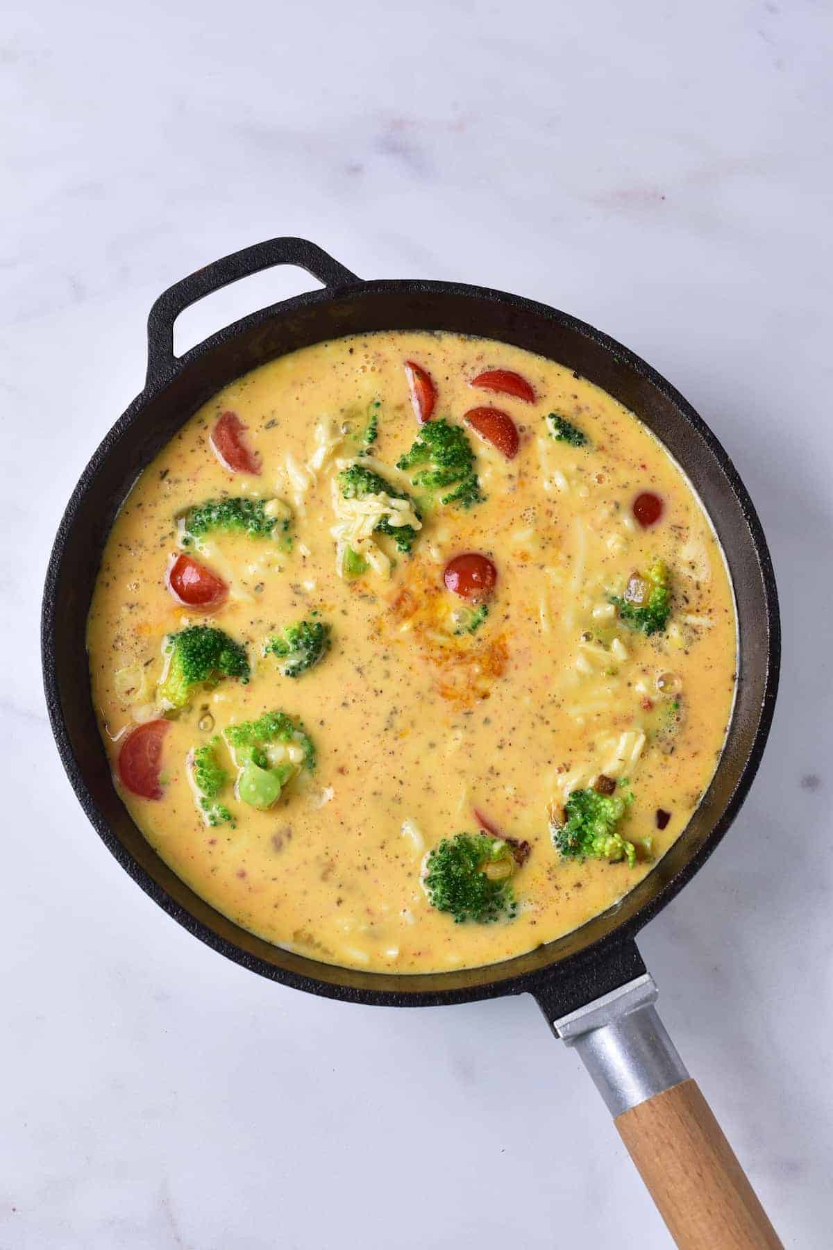 A black skillet filled with an uncooked egg mixture, broccoli, cherry tomatoes, and shredded cheese on a white surface.