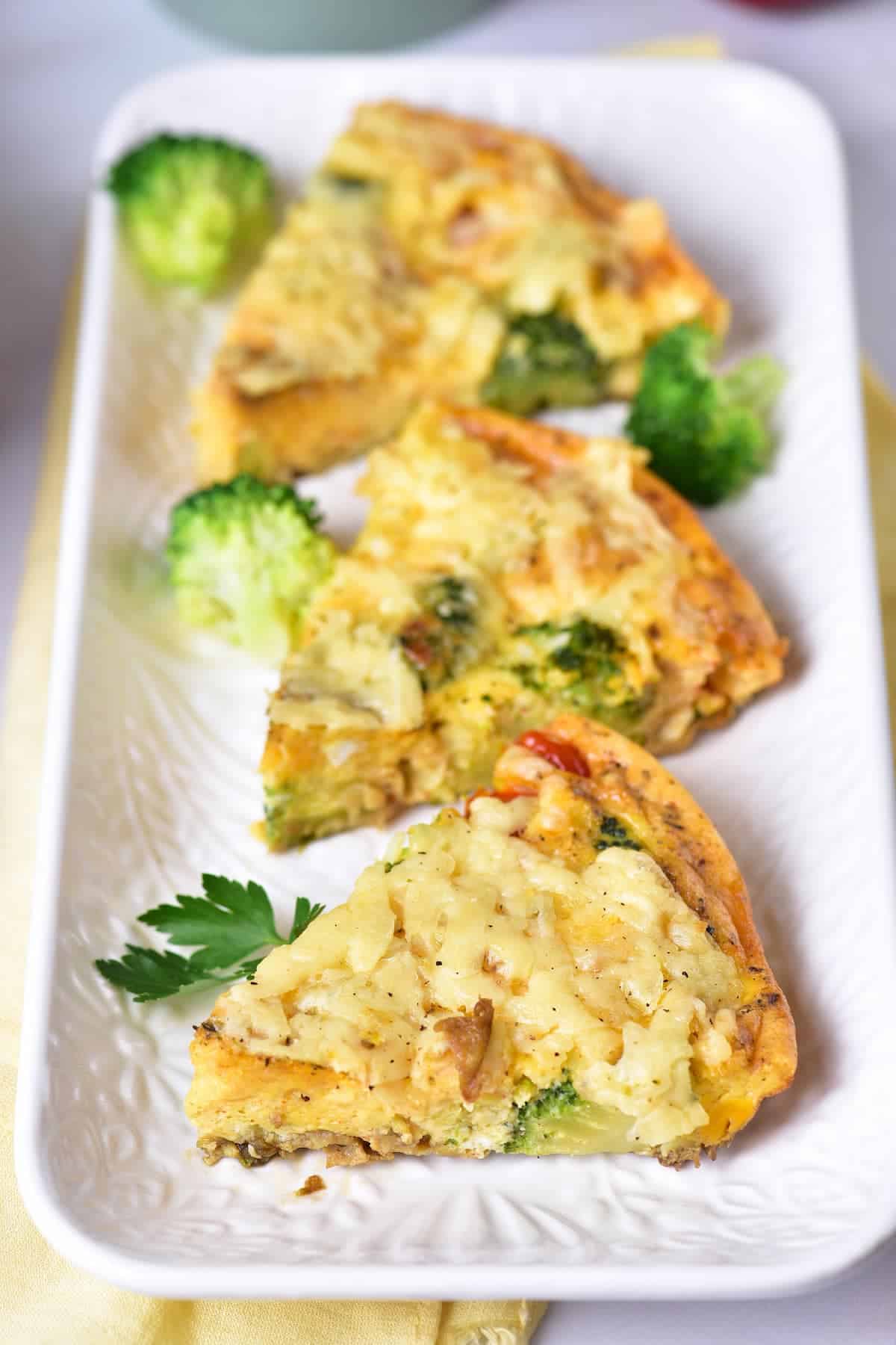 Three slices of vegetable frittata, garnished with broccoli florets and a sprig of parsley, are arranged on a rectangular white plate.