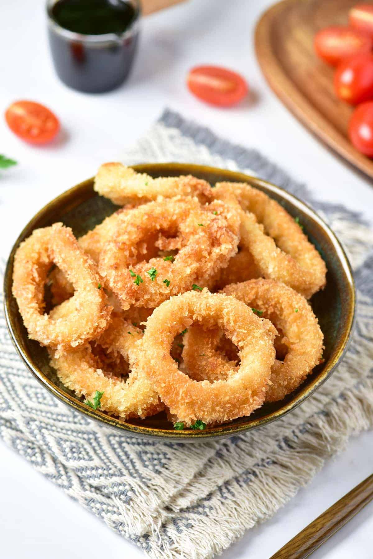 On a wooden plate nearby, slices of tomato and a cup of coffee sit beside a bowl of crispy calamari fries with sliced lemons and a hint of sauce on one piece, all set on a mat.