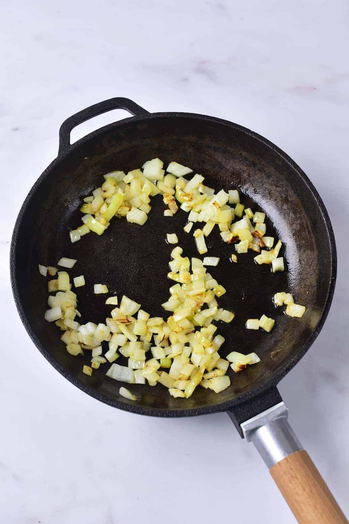 Chopped onions being sautéed in a black frying pan with a wooden handle on a white surface.