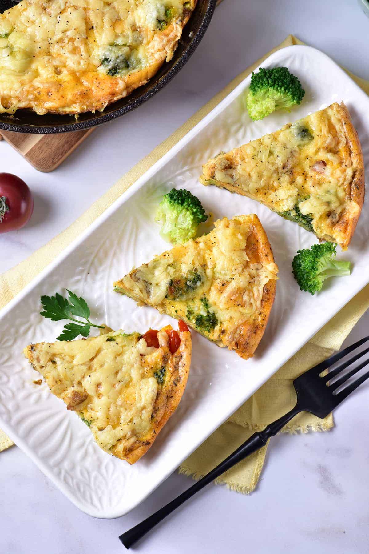 Three slices of vegetable quiche are arranged on a rectangular white plate with broccoli florets and parsley garnish, next to a black fork and a yellow napkin.