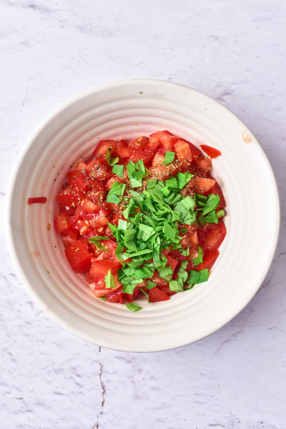 A white bowl containing chopped tomatoes, fresh chopped basil, and seasoning, placed on a light marble surface.