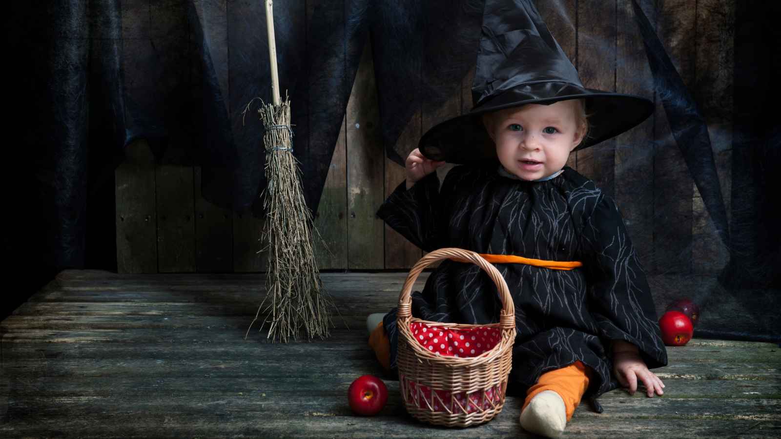 A young child dressed as a witch sits on a wooden floor with a broom, a wicker basket, and apples around them, against a rustic wooden backdrop.