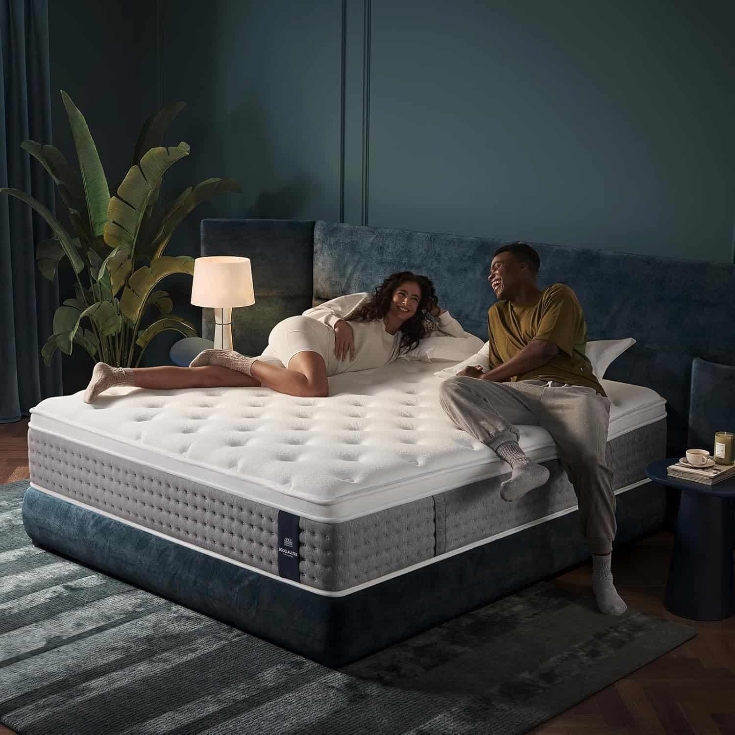 A woman and a man sit smiling on a large bed with a white mattress in a modern, dark-colored bedroom with a plant, a lamp, and a rug.