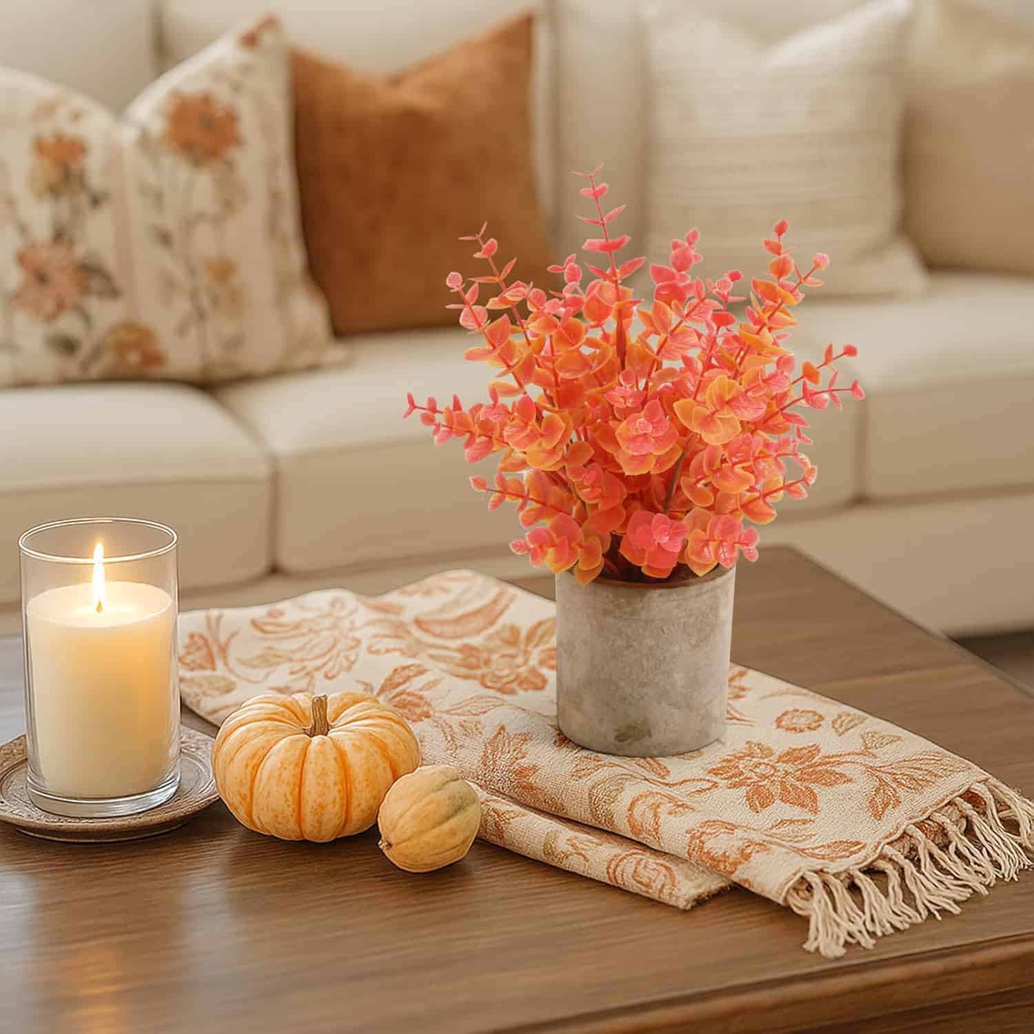 A lit candle, two small pumpkins, and a potted orange plant are arranged on a patterned cloth atop a wooden coffee table in a living room.