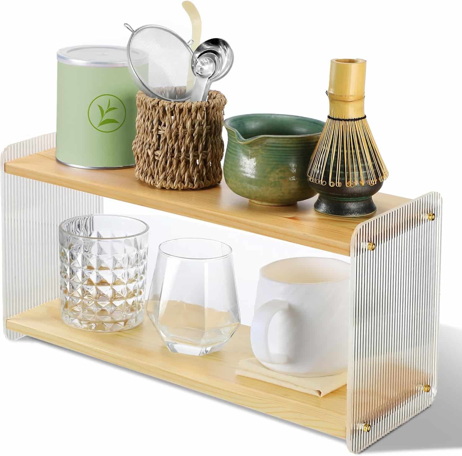 A small two-tiered wooden and acrylic shelf holding a tin, utensils, whisk, cups, glasses, and a woven container, used for organizing kitchen or tea items.