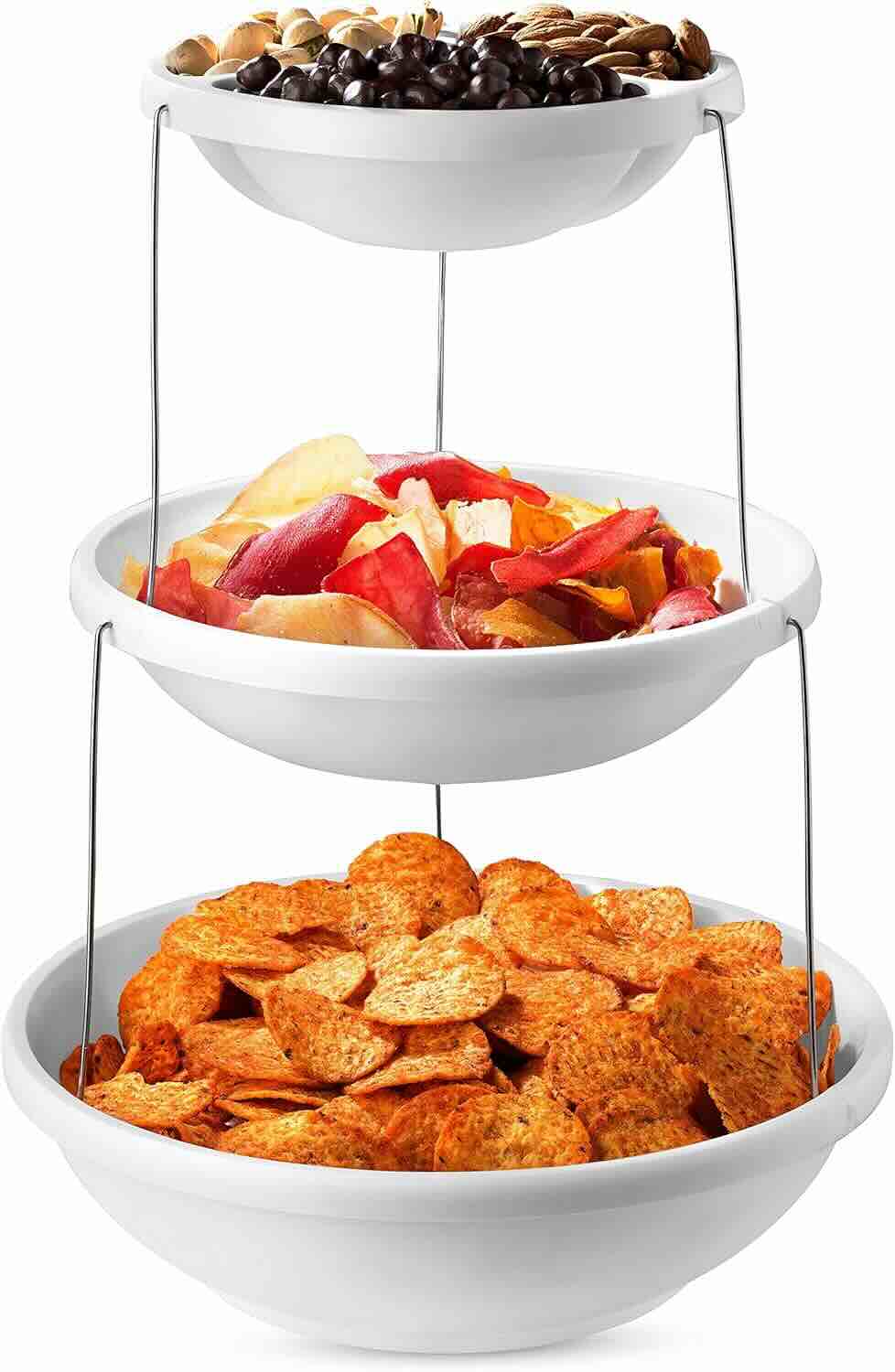 Three-tiered white serving tray filled with snacks: pistachios and chocolates on top, mixed vegetable chips in the middle, and round orange chips on the bottom.
