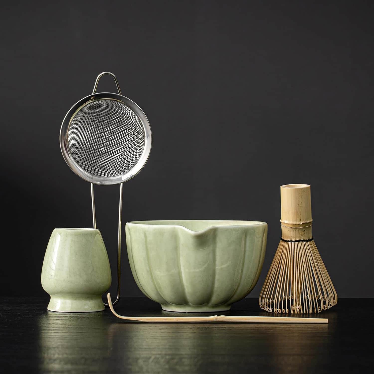 A traditional matcha tea set with a ceramic bowl, whisk holder, whisk, sifter, and bowl, arranged against a dark background.