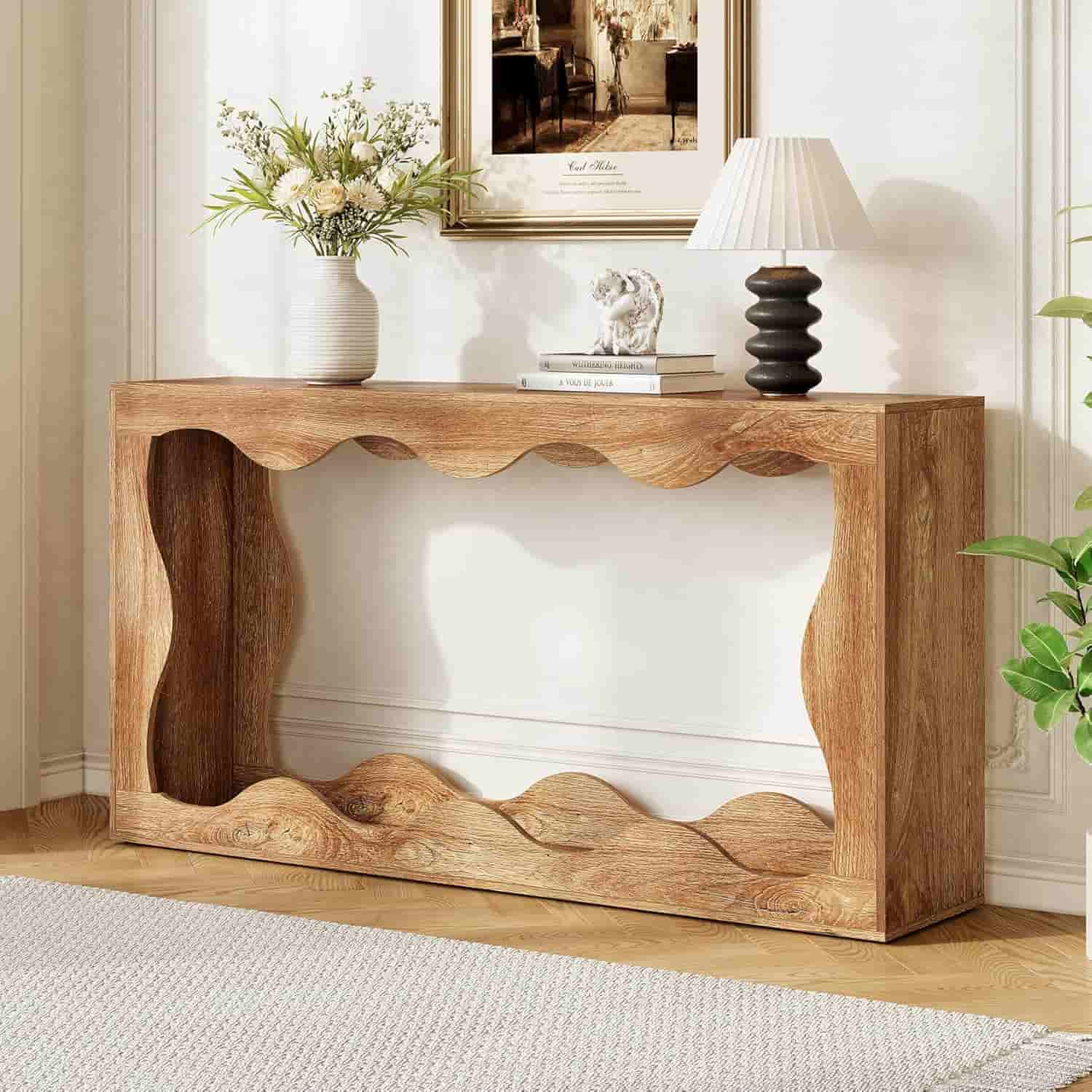 A wooden console table with wavy cut-out design stands against a wall, topped with a vase of flowers, a lamp, and a decorative sculpture. Framed art hangs above.
