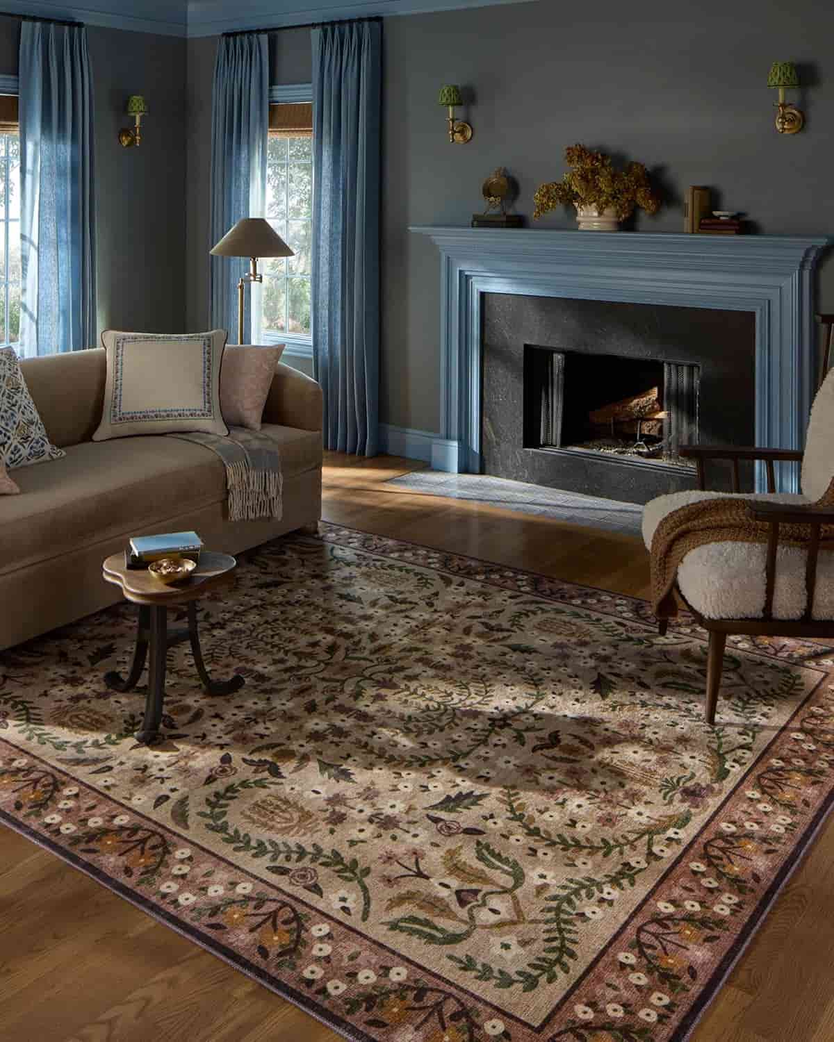 A living room with a beige sofa, an armchair, a floral-patterned rug, blue curtains, a dark fireplace, and a side table with a cup, all in natural light.