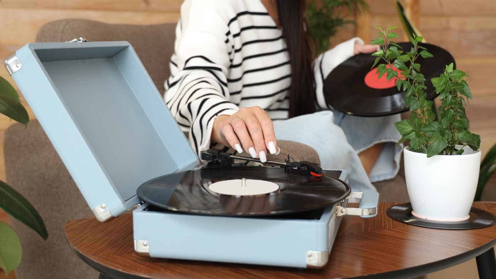 A person places a vinyl record on a blue portable turntable, with another record and a potted plant on the table nearby.