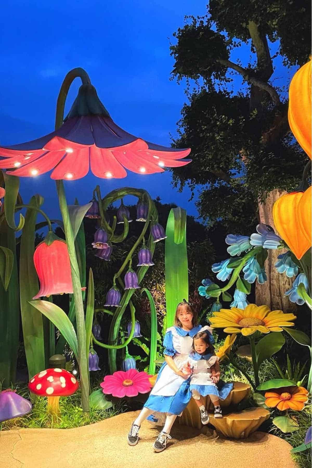 Two children dressed as Alice in Wonderland sit on a giant mushroom amid oversized, colorful flowers and plants in a whimsical, fantasy-themed setting.