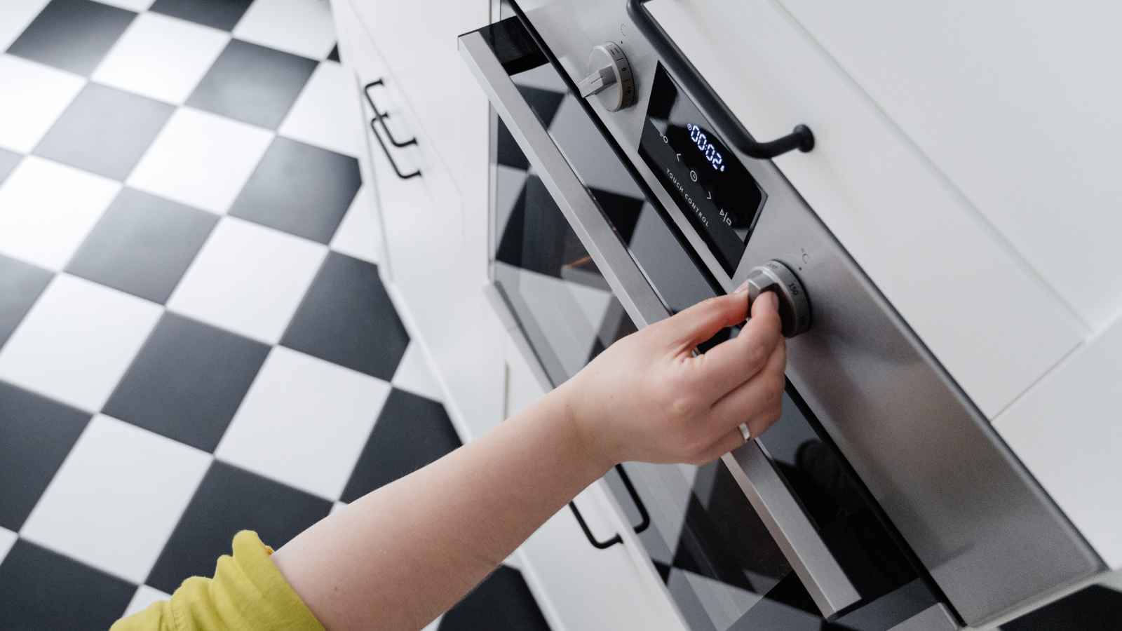 A person in a yellow sleeve adjusts the temperature knob on a stainless steel oven in a kitchen with a black and white checkered floor.