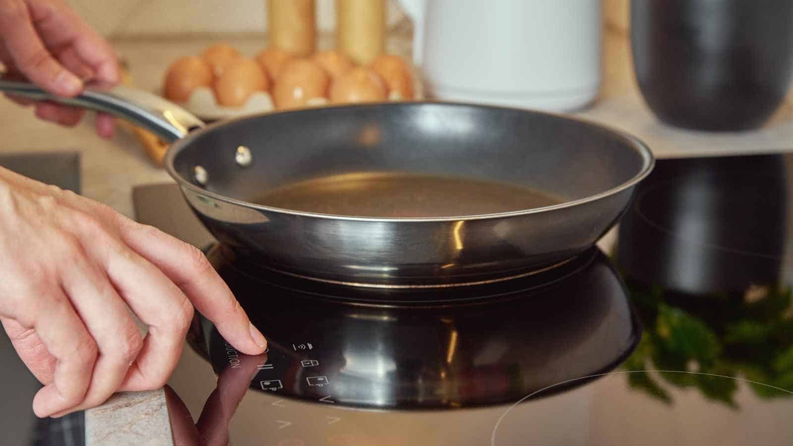 A person turns on an induction cooktop with a frying pan placed on it; eggs and kitchen items are visible in the background.