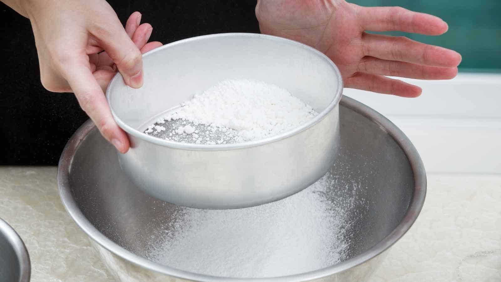 Hands sifting white powder, likely flour or sugar, through a metal sieve into a mixing bowl on a light countertop.