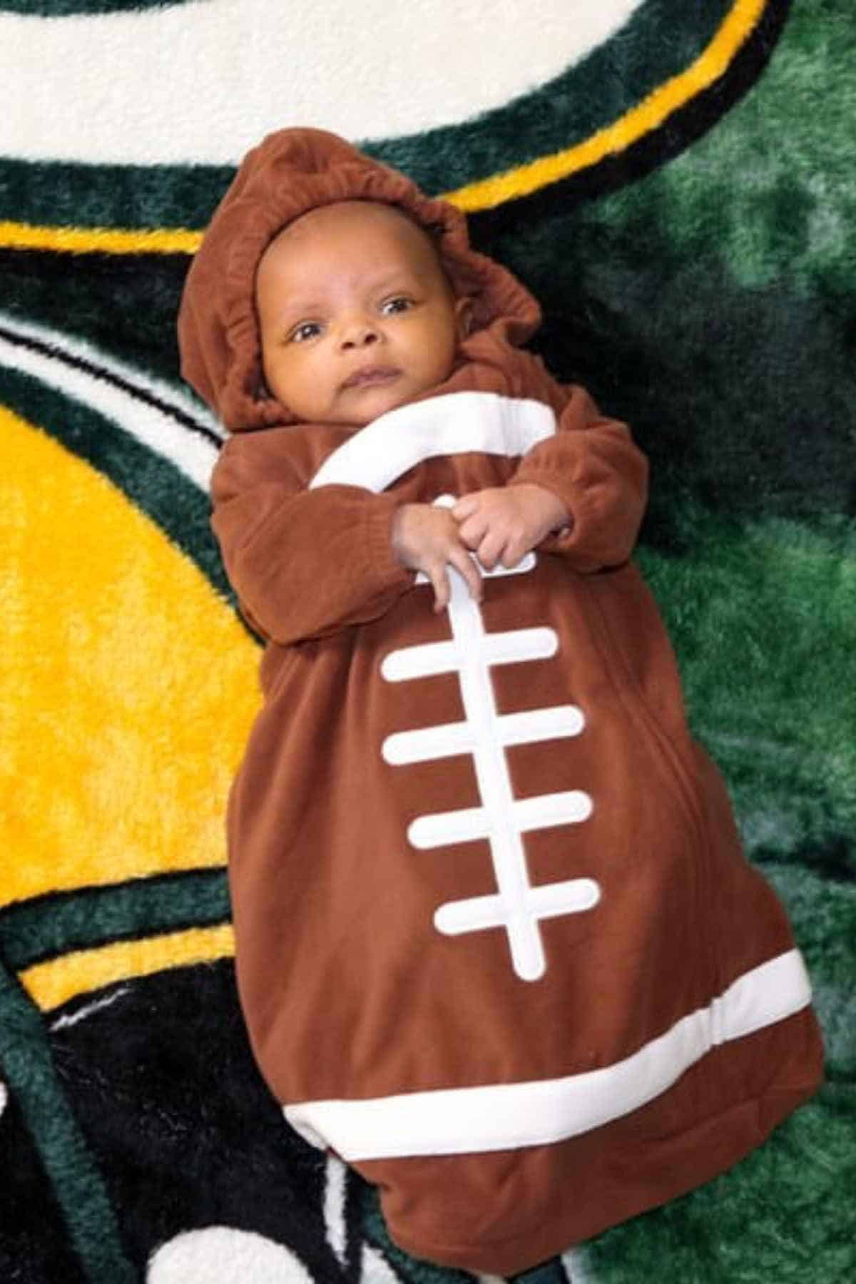 A baby dressed in a brown football-shaped outfit lies on a blanket with green, yellow, and white patterns.