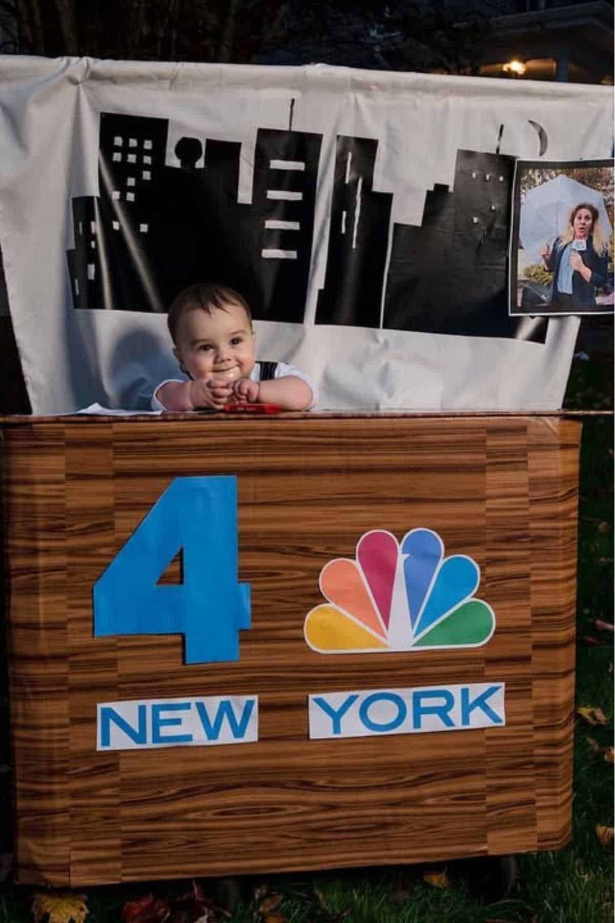 A baby sits behind a homemade news desk with a "4 New York" NBC logo, a cityscape backdrop, and a framed photo of a weather anchor on the side.