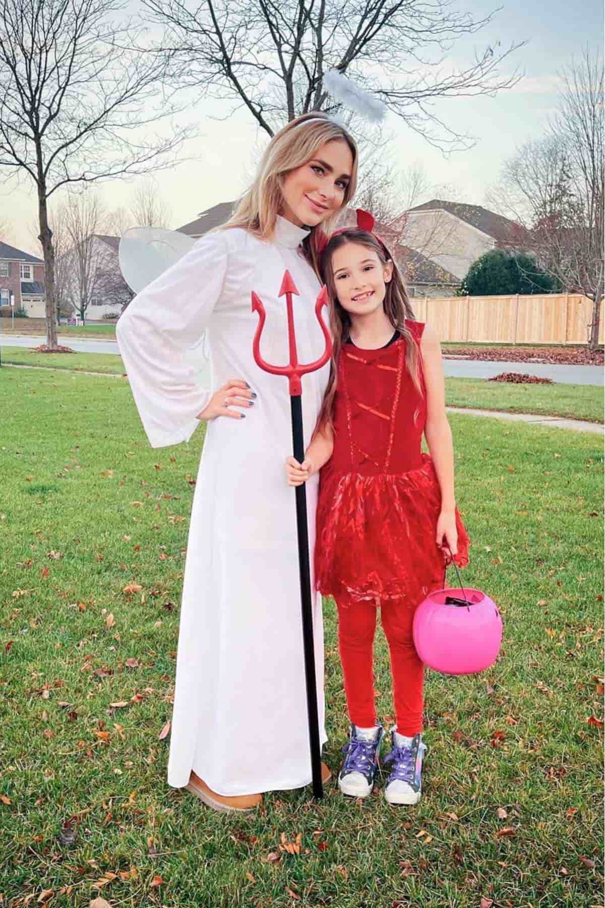An adult dressed as an angel and a child dressed as a devil stand together on grass, posing for a photo. The child holds a pink pumpkin-shaped bucket.