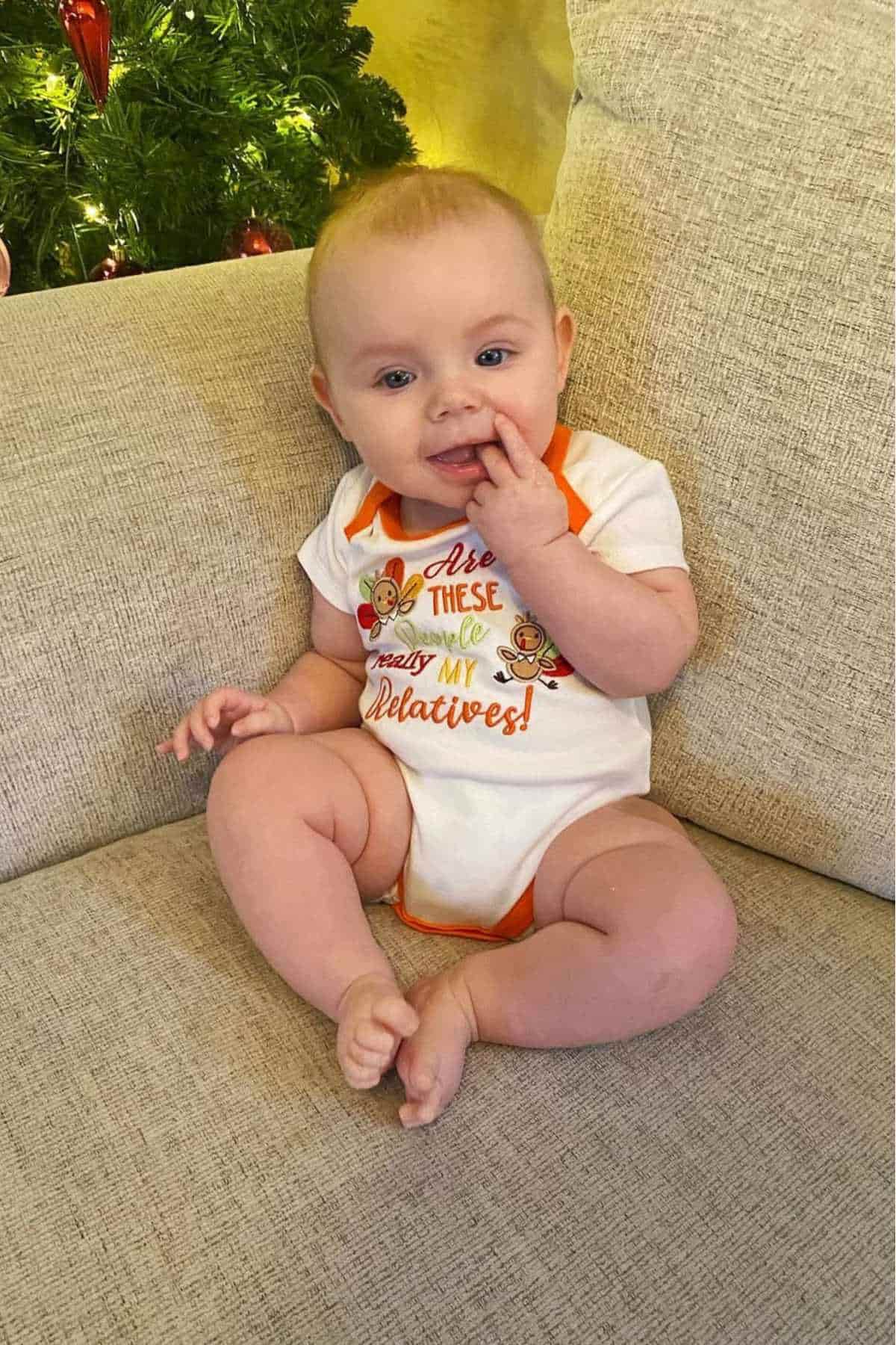 A baby sits on a beige couch with a hand near their mouth, wearing a festive onesie. A decorated Christmas tree is partially visible in the background.