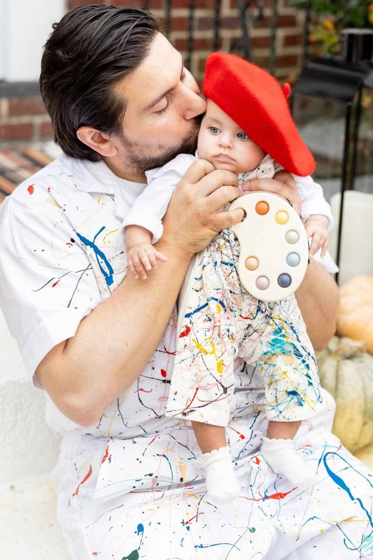 An adult wearing paint-splattered clothes kisses a baby dressed in similar paint-splattered clothes, a red beret, and a paint palette accessory.