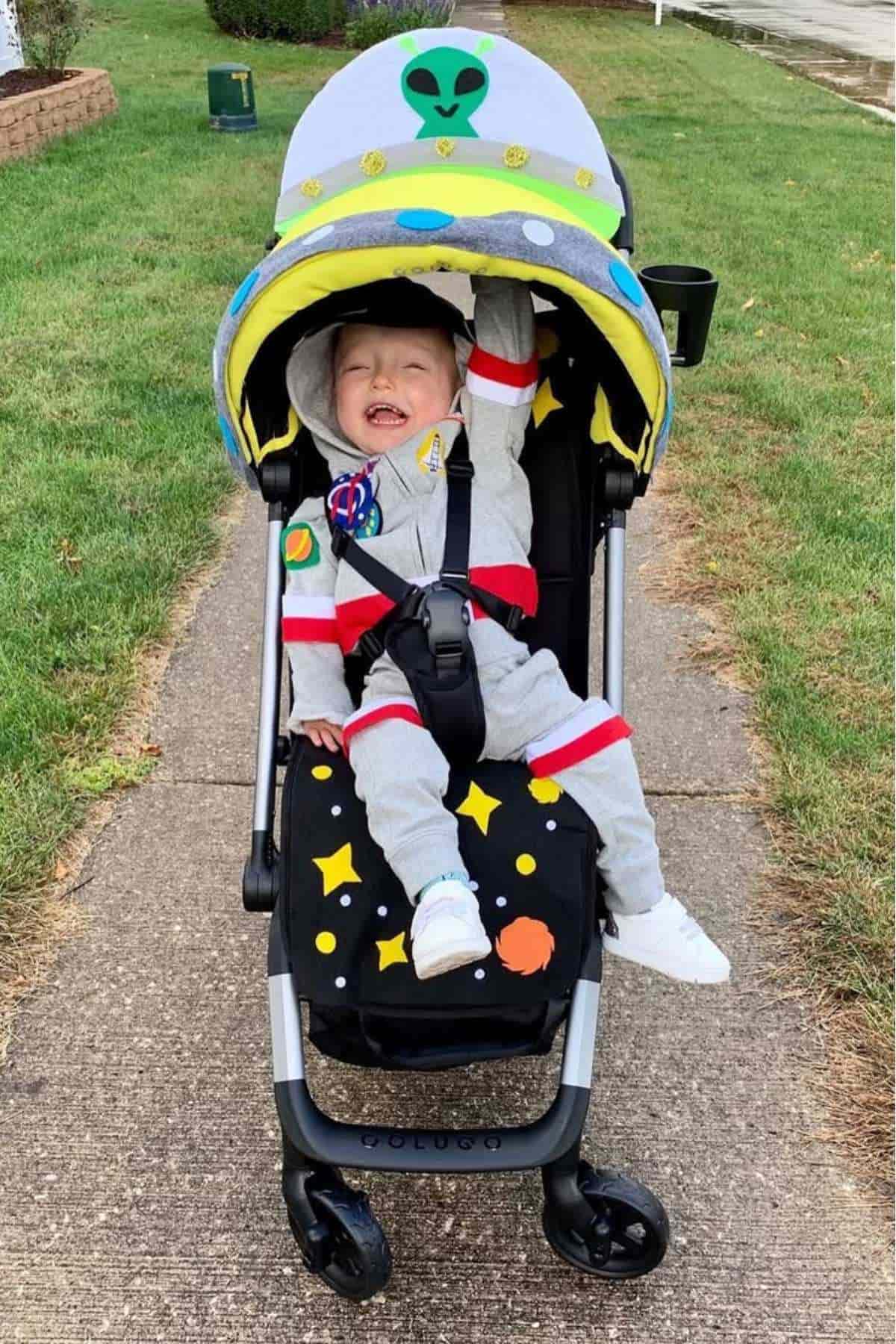Smiling child in a stroller decorated with an alien and space theme, wearing a gray spacesuit costume on a sidewalk.