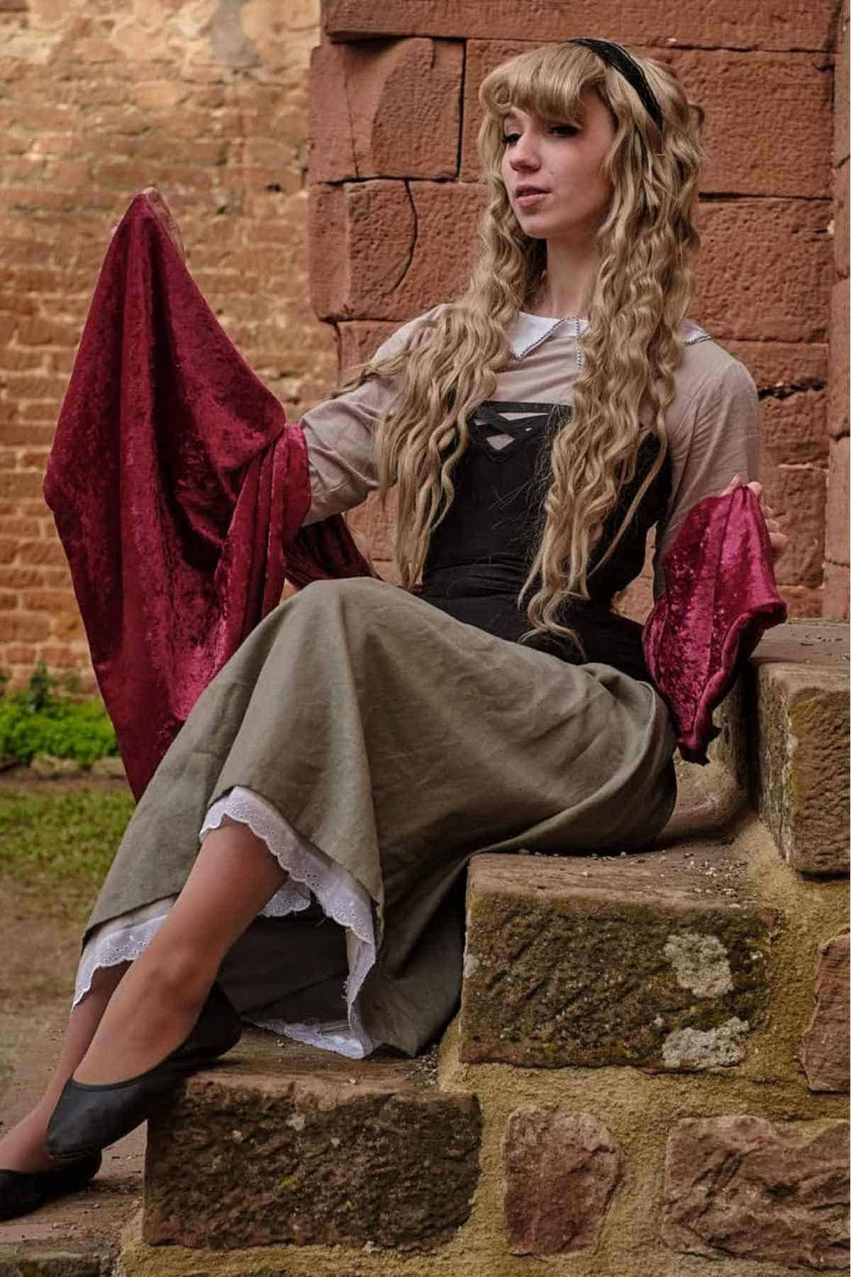 A woman in a medieval-style dress with long, wavy hair sits on stone steps near a brick wall, holding red fabric in both hands.
