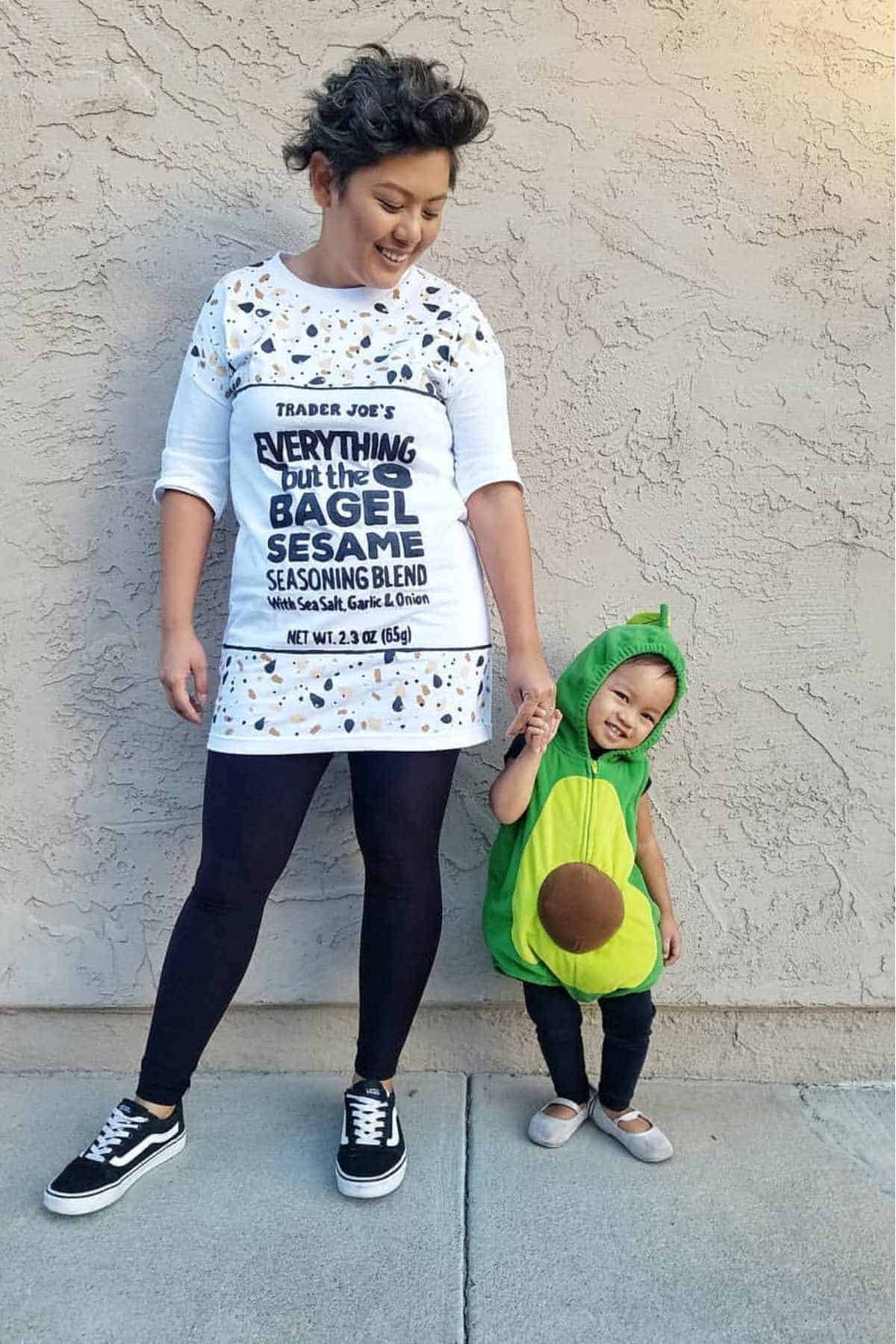 An adult in a bagel seasoning costume stands next to a child wearing an avocado costume; both are smiling and holding hands against a beige wall.