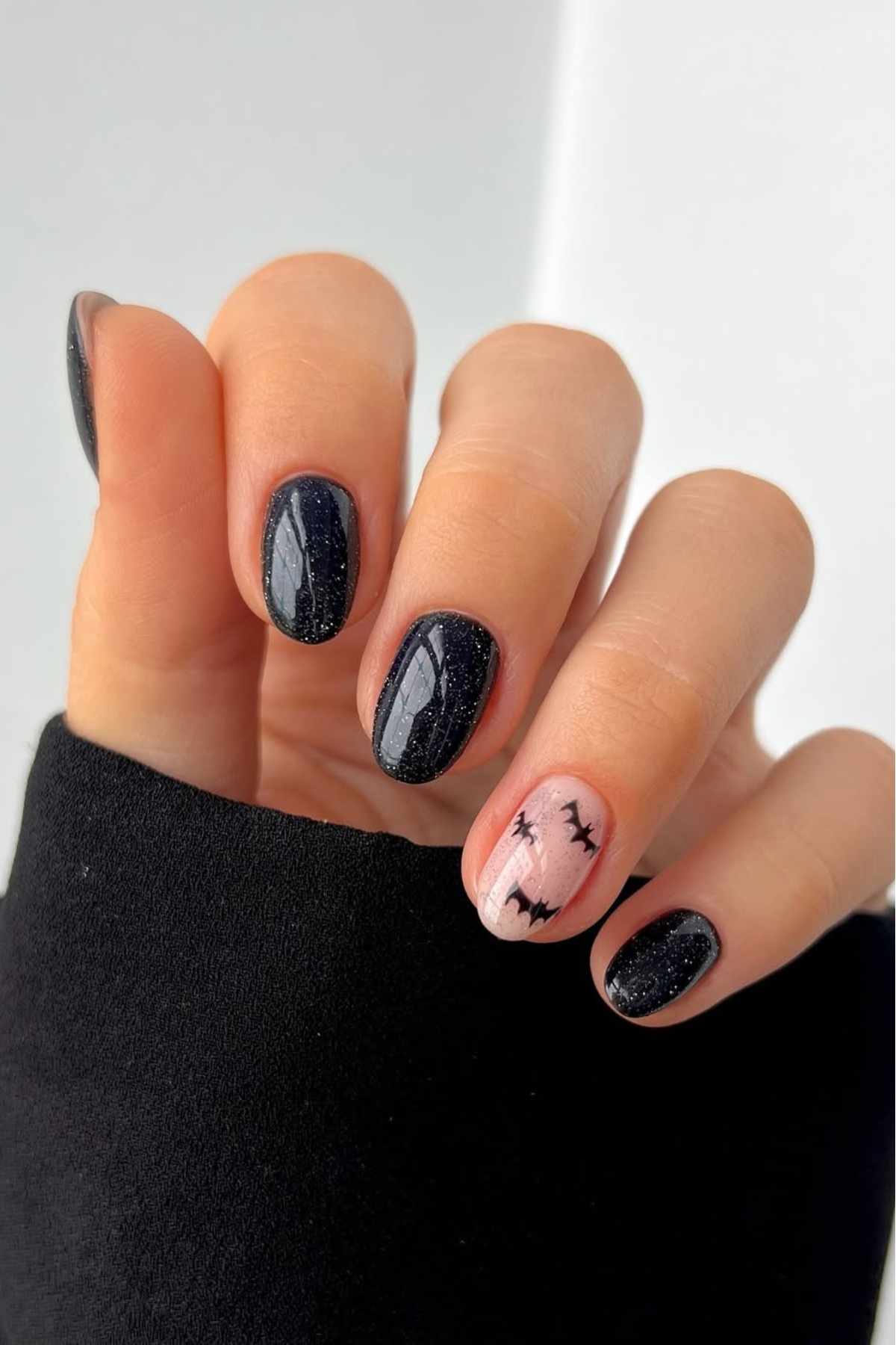 A hand with short nails painted black, except for the ring finger, which has a pink base with a bat design.
