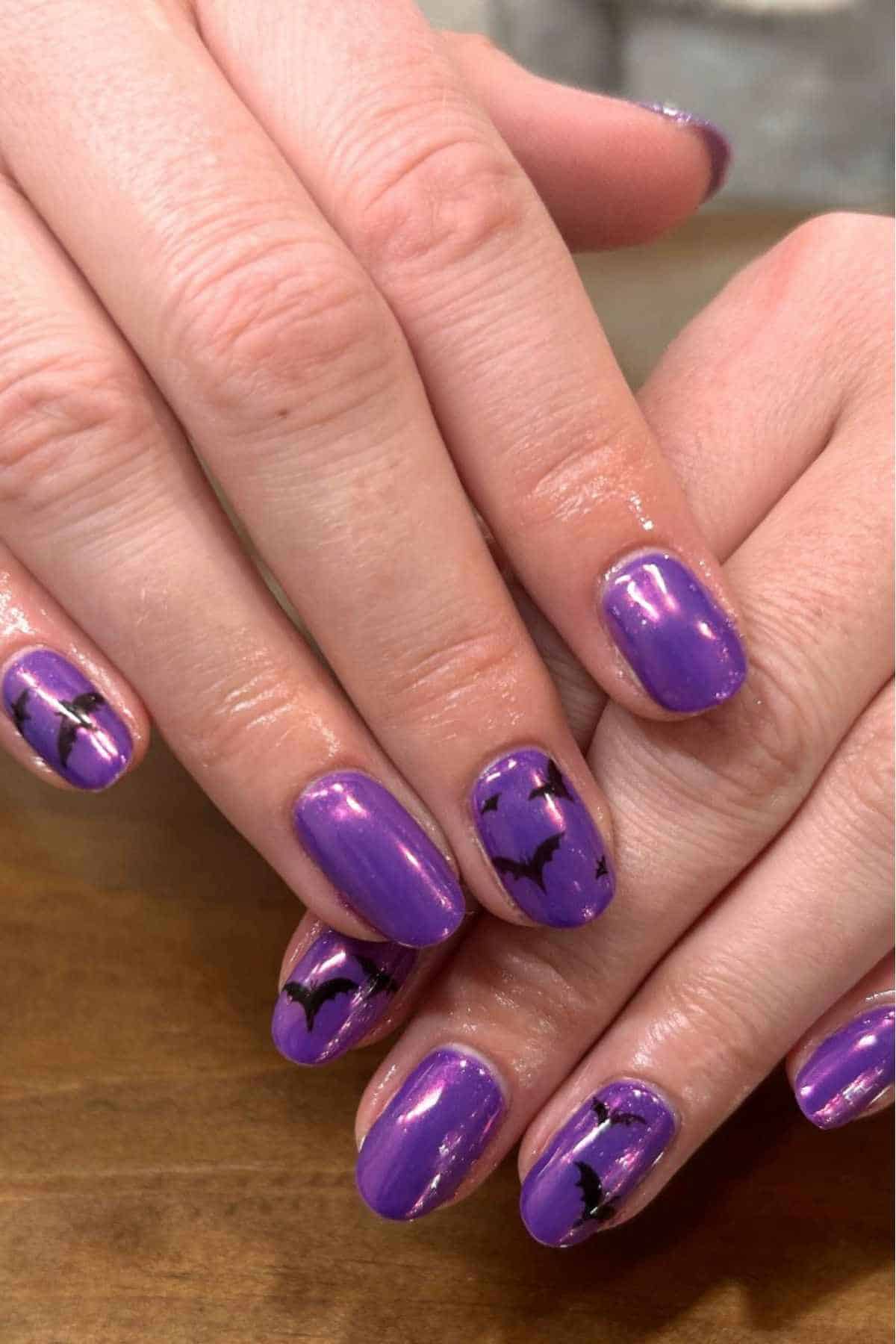 Hands with purple nail polish featuring black bat designs on several nails, resting on a wooden surface.