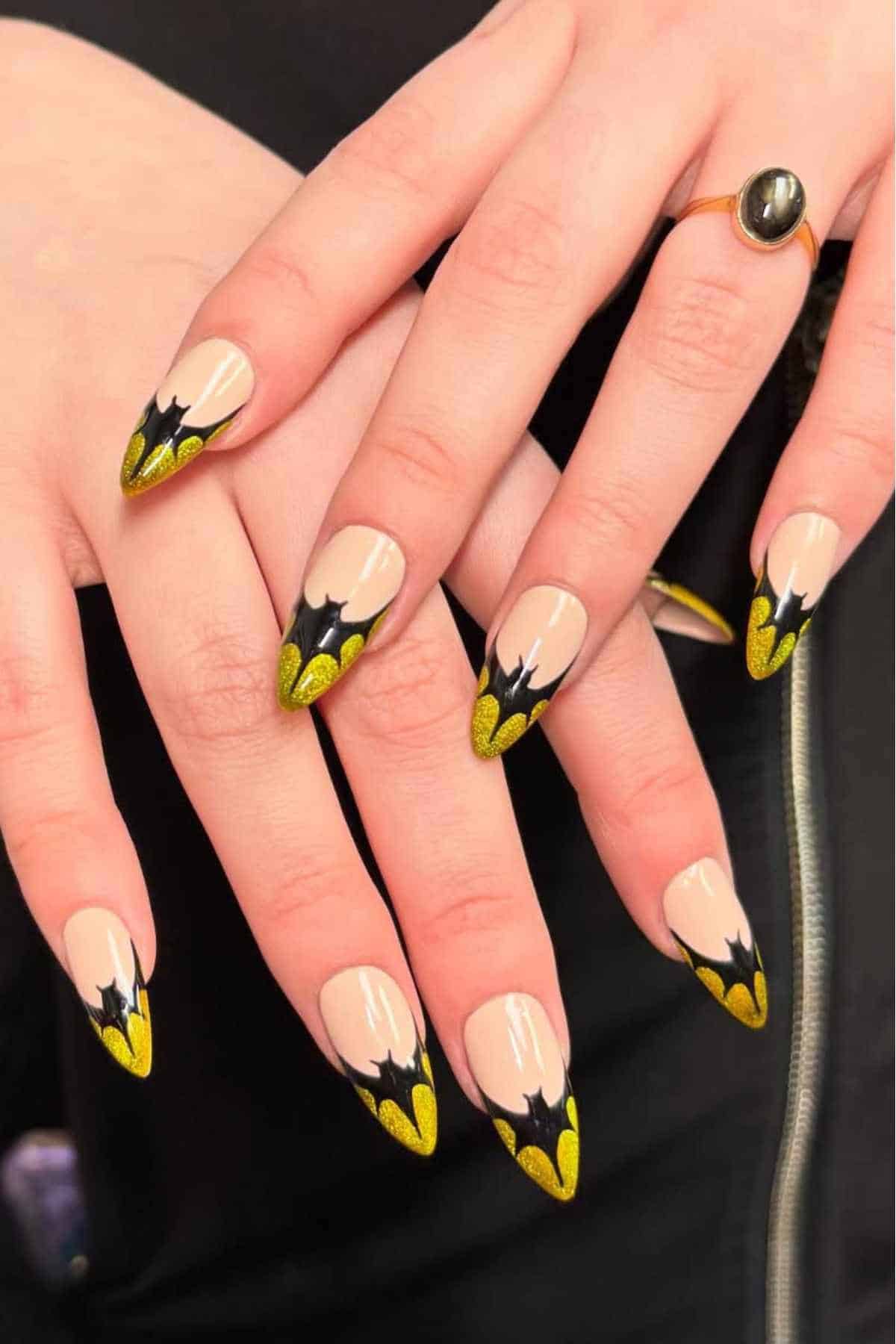 Hands with long, almond-shaped nails featuring a nude base and yellow and black bat wing nail art tips.