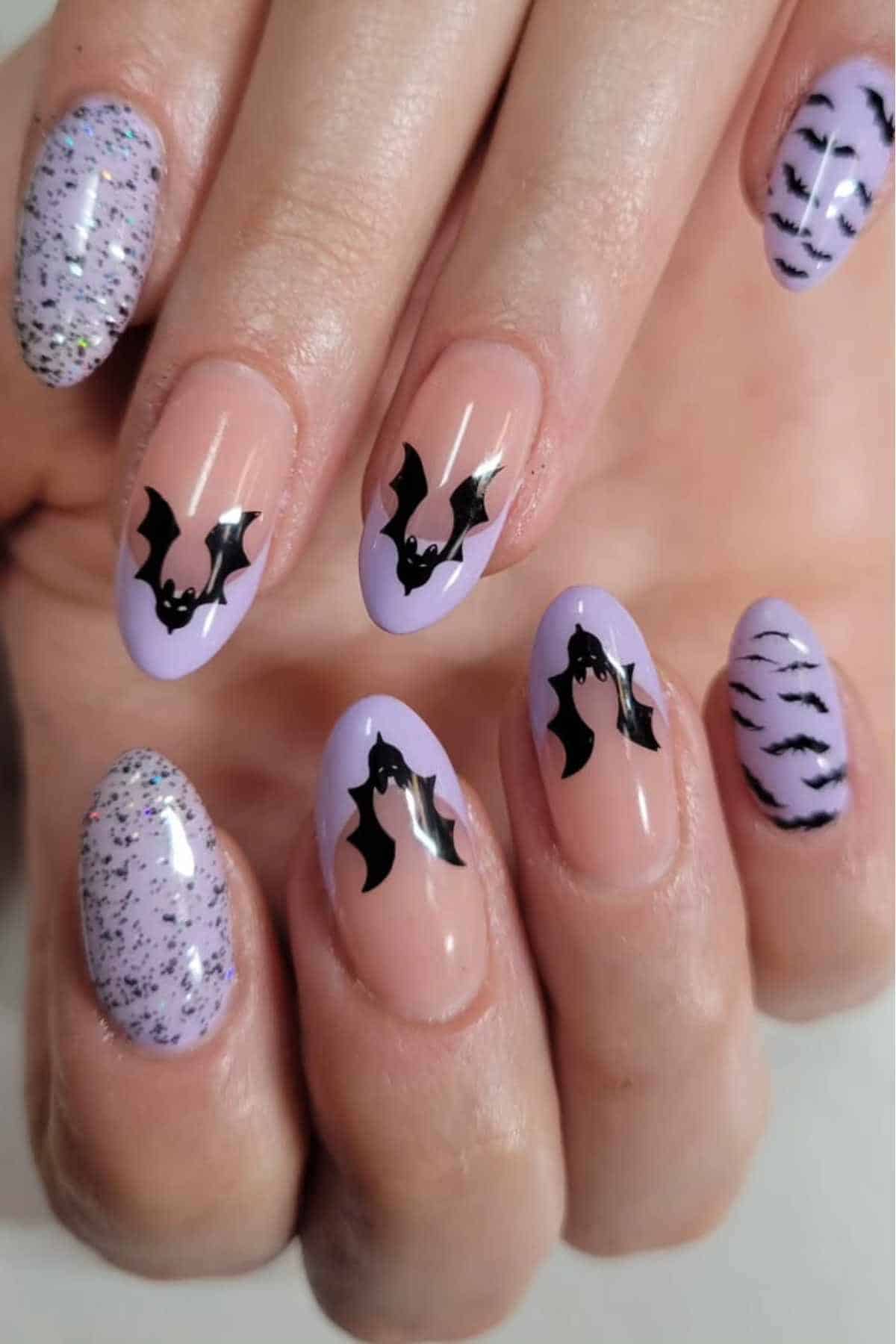 A hand with long, almond-shaped nails painted in lavender shades, featuring black bat designs, glitter, and wavy line patterns.