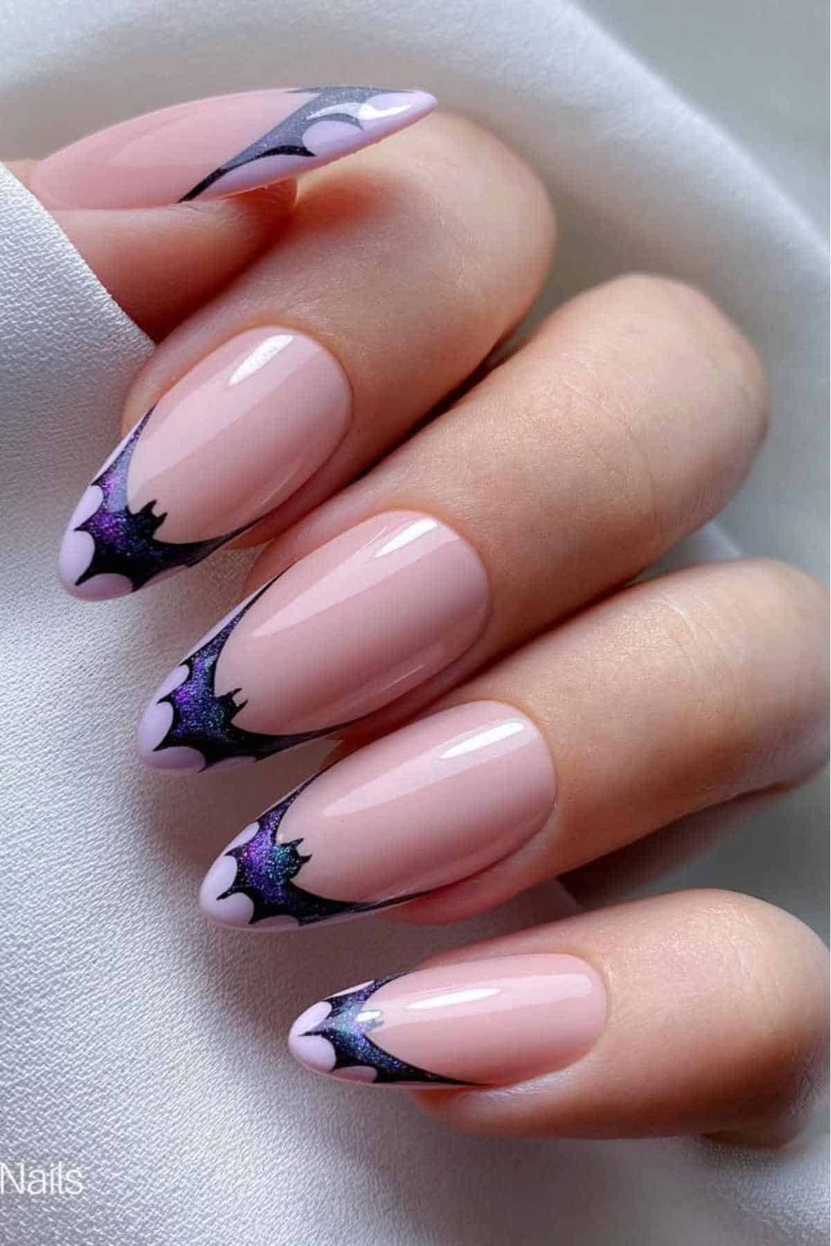 A hand with almond-shaped nails painted nude pink, featuring black and purple bat-shaped tips on a white background.