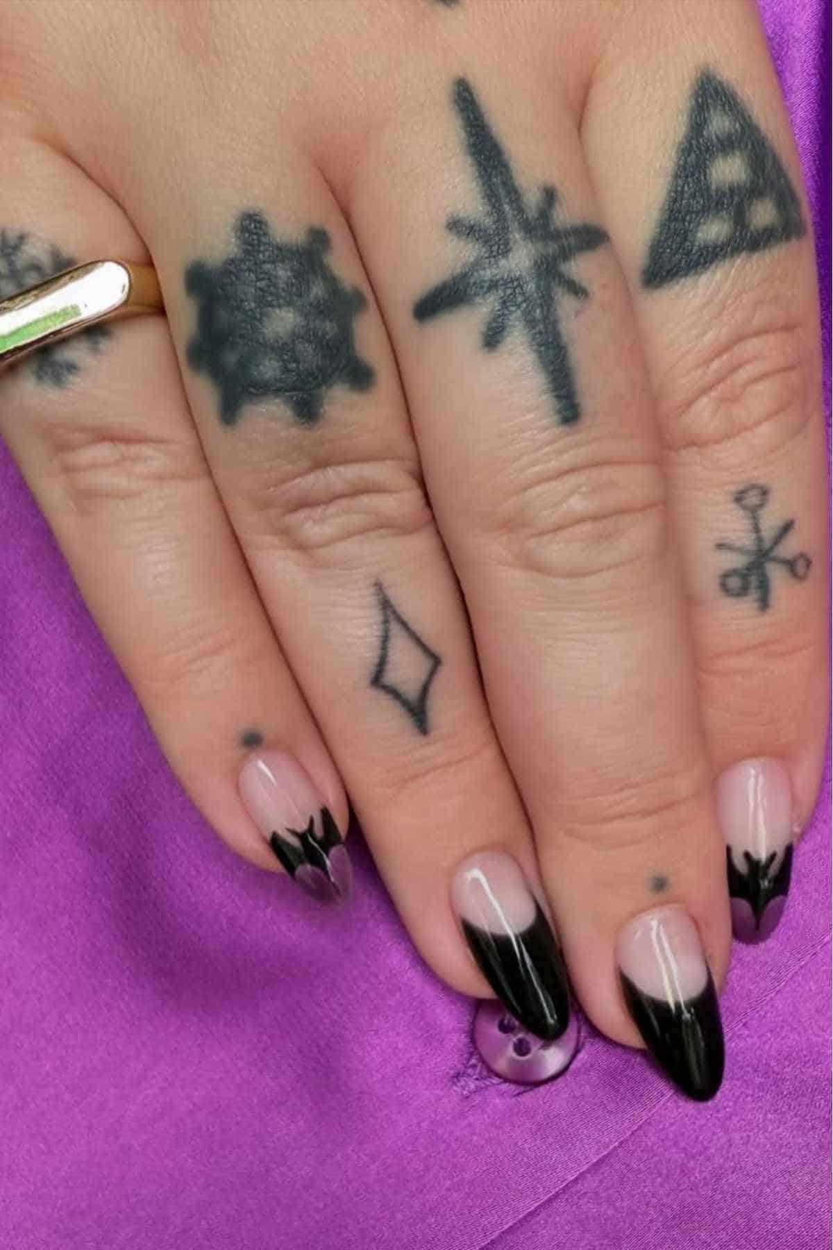 A hand with a black tattoos and long almond-shaped nails featuring a black French tip design rests on a purple fabric.
