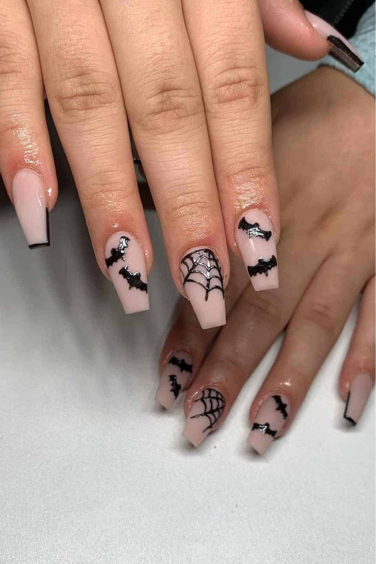 Close-up of hands with nude coffin-shaped nails featuring black bat and spiderweb Halloween nail art designs.