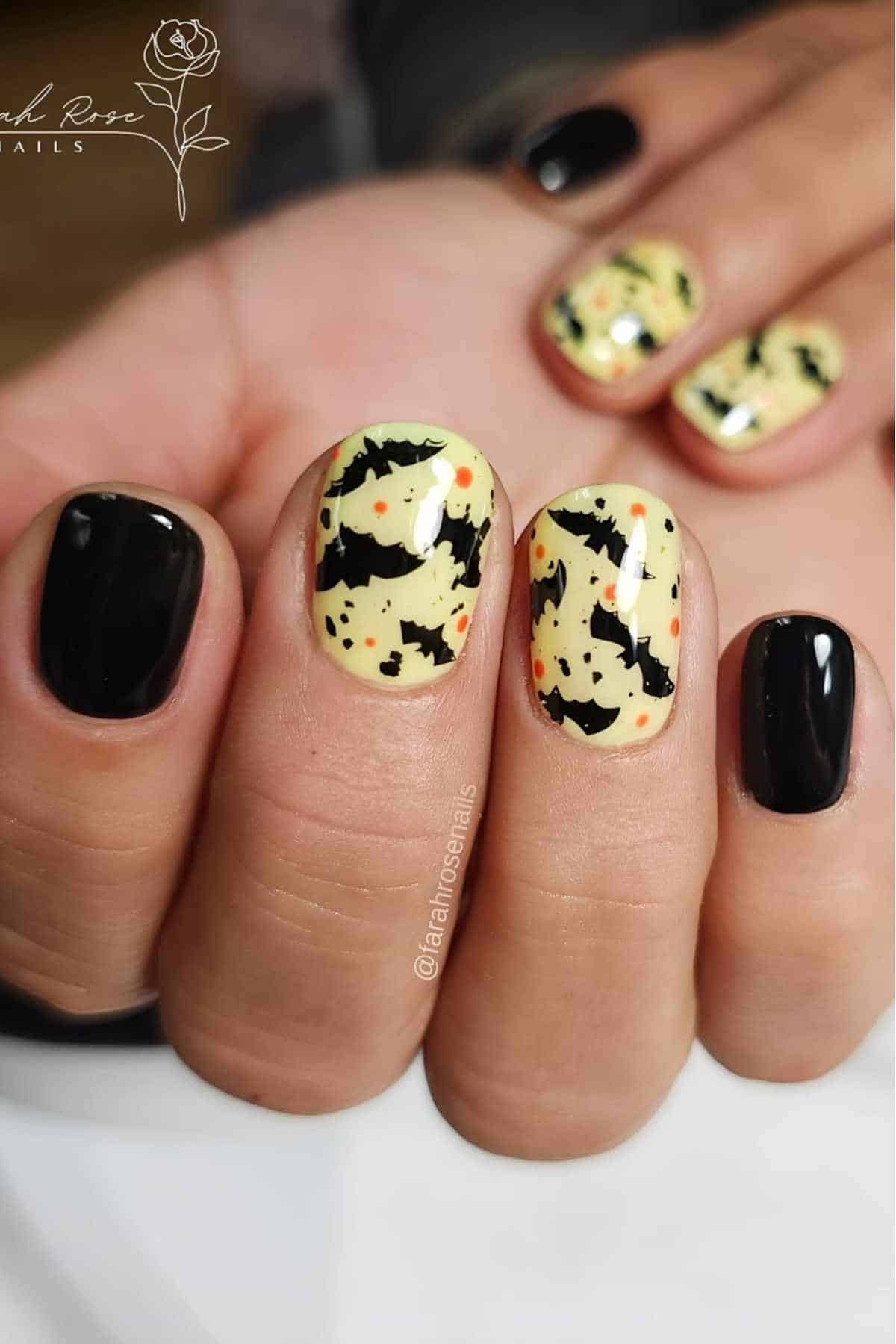Close-up of fingernails with Halloween-themed nail art; three nails have black bats and orange dots on a pale yellow base, others are painted solid black.