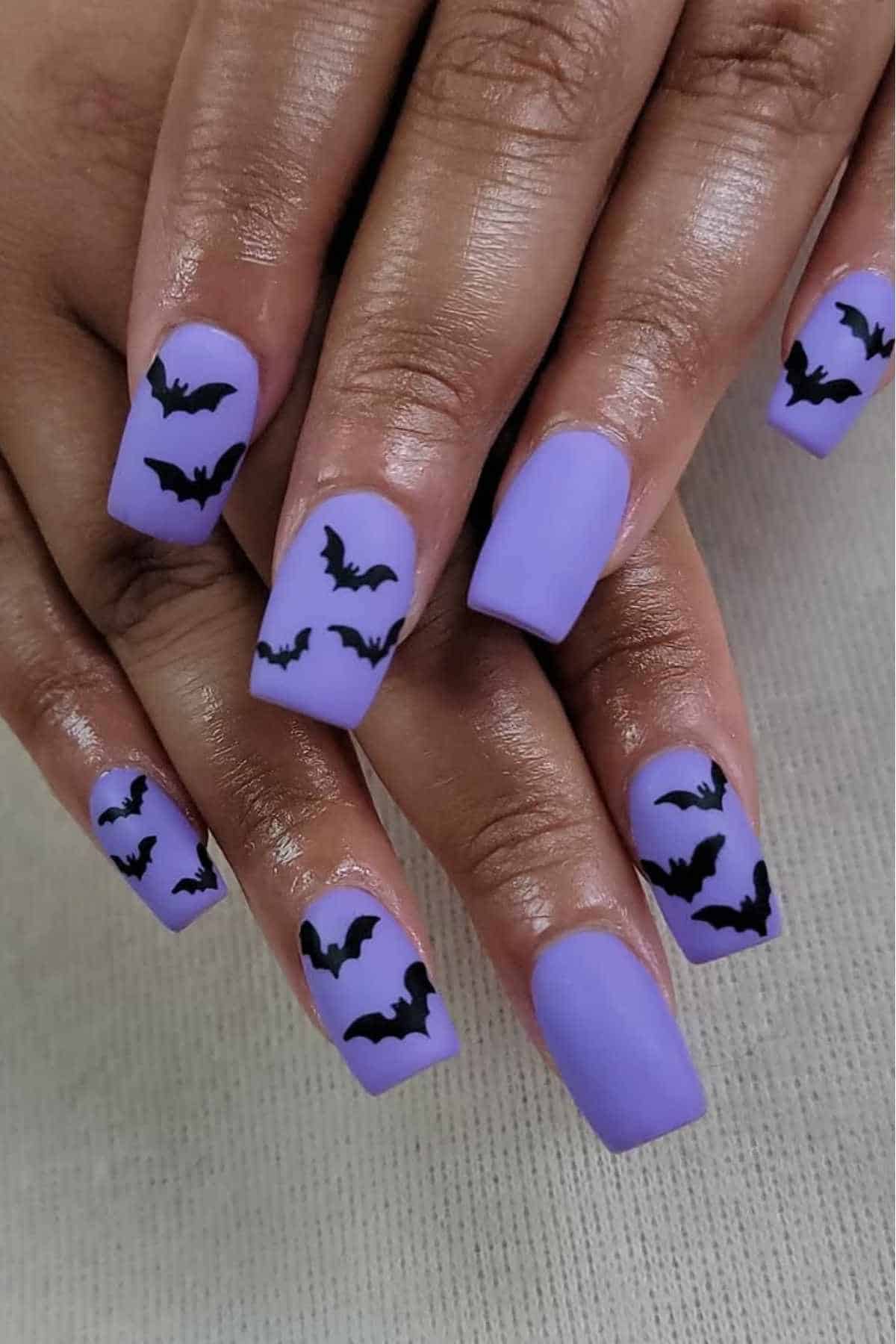 Hands with light purple square-shaped nails featuring black bat silhouettes as nail art.