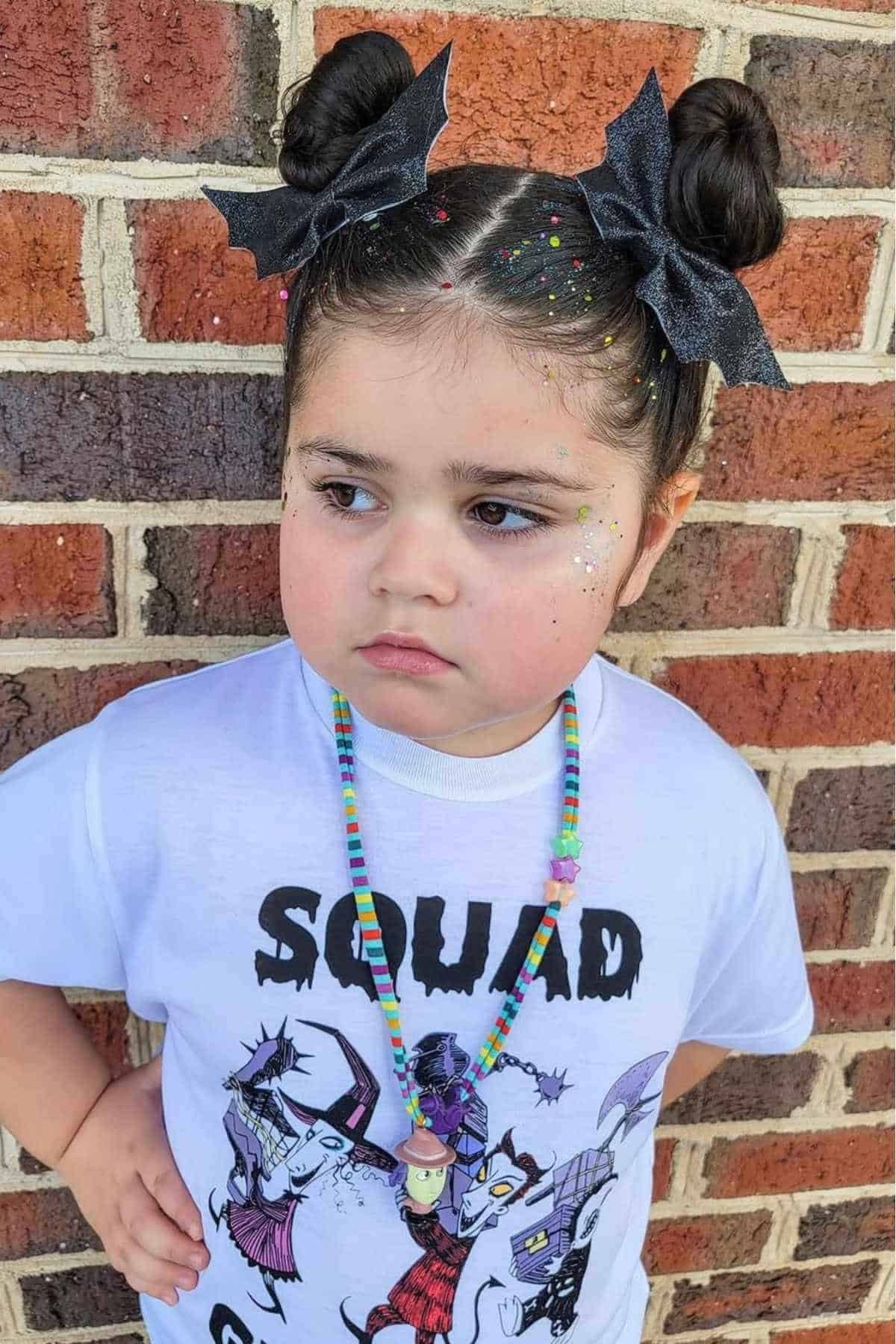 Young child standing against a brick wall, wearing a "Squad" graphic t-shirt, colorful beaded necklace, glitter on face, and hair styled in two buns with large black bows.