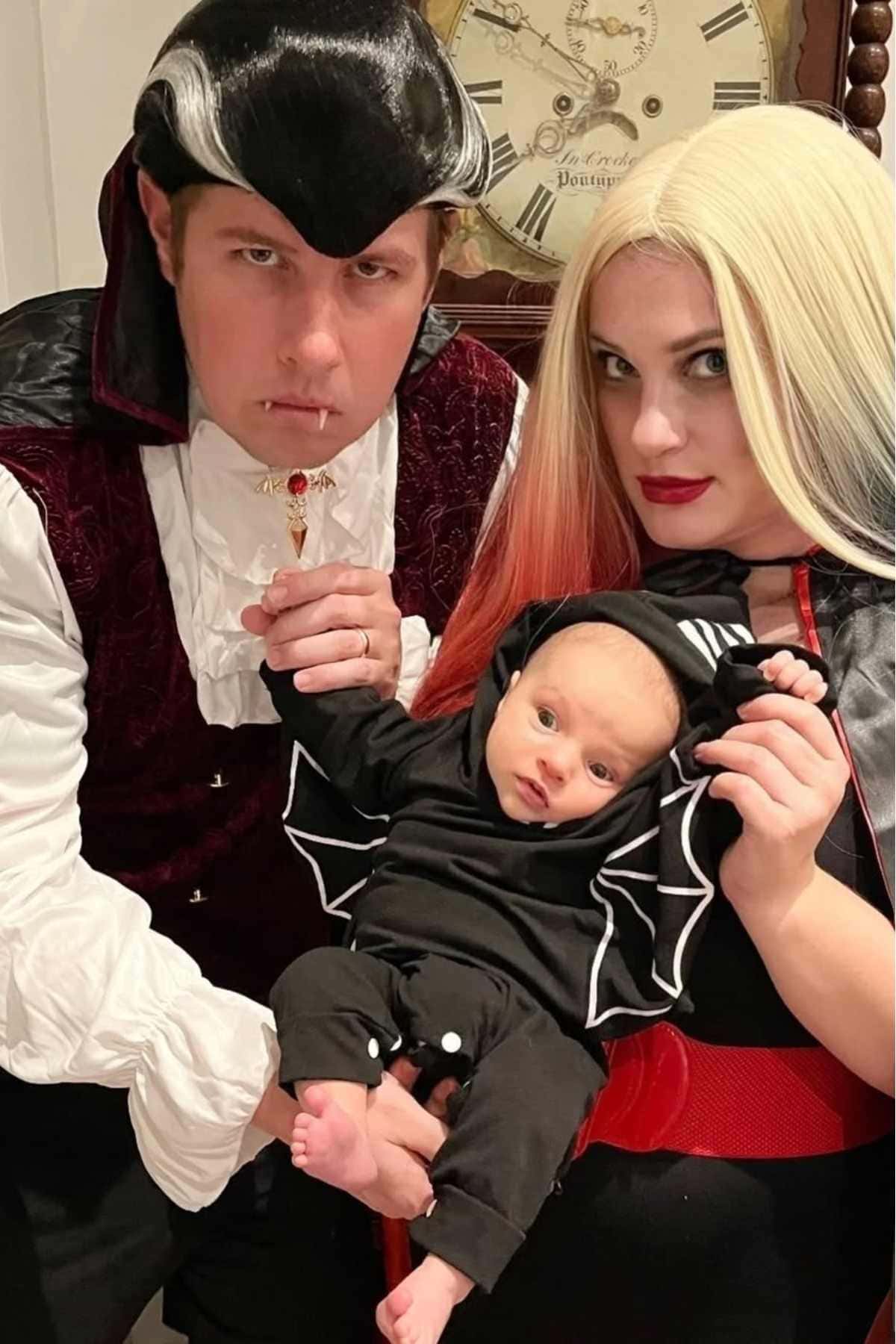 Three people in vampire-themed costumes pose together; an adult male, an adult female with black and blonde hair, and a baby in a black bat outfit. A large clock is in the background.