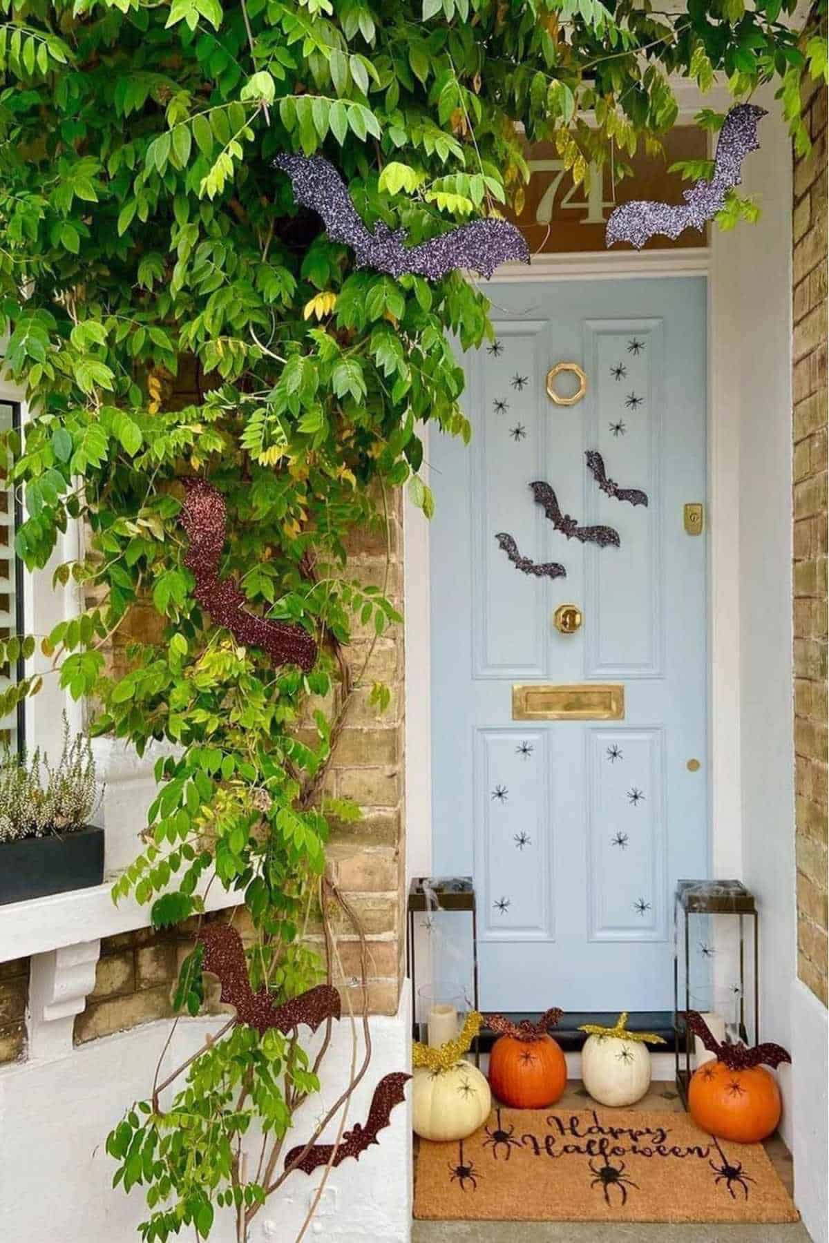A front door decorated with black bat cutouts, pumpkins, two lanterns, and a "Happy Halloween" doormat, with green vines framing the entrance.