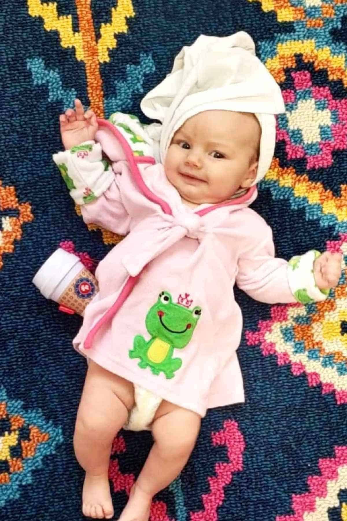 Baby lying on a colorful patterned rug, wearing a pink robe with a frog design, a towel on their head, and a disposable coffee cup beside them.