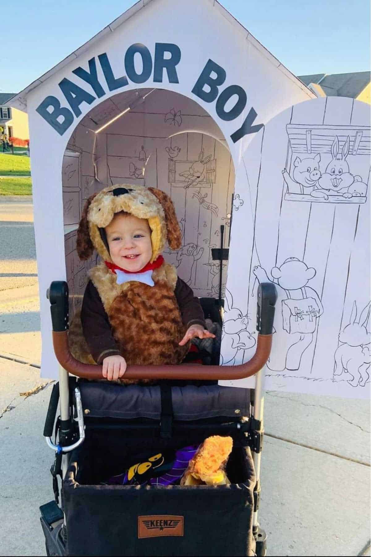 A young child in a dog costume sits in a stroller decorated as a doghouse with "Baylor Boy" written above the entrance.