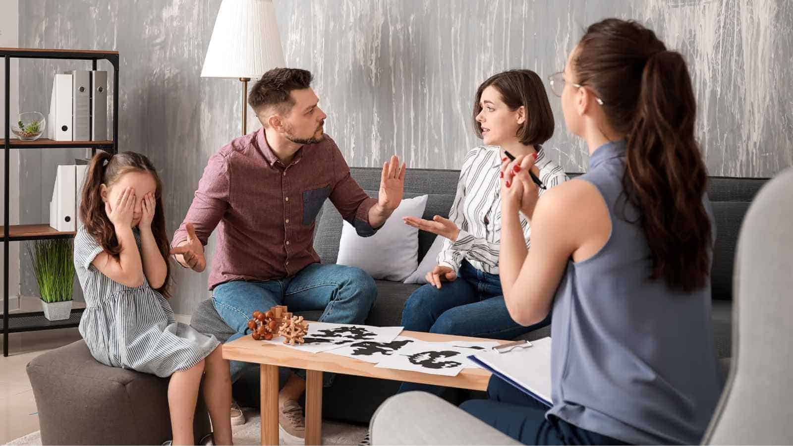 A man and woman argue while sitting on a couch during a therapy session; a young girl covers her face, and a therapist observes with a clipboard.