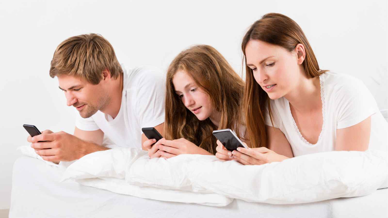Three people lying on a bed, each looking at their own smartphone. They appear focused on their devices and are dressed in white shirts.