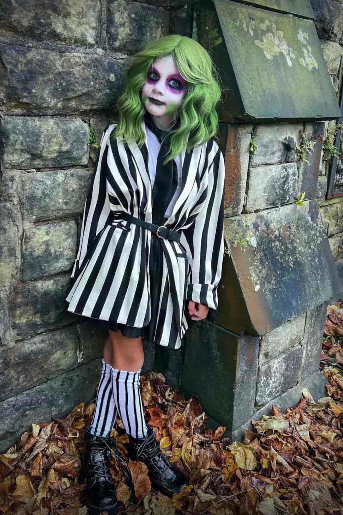 Child in a black and white striped outfit with green hair and dramatic makeup stands against a stone wall covered in moss and fallen leaves.