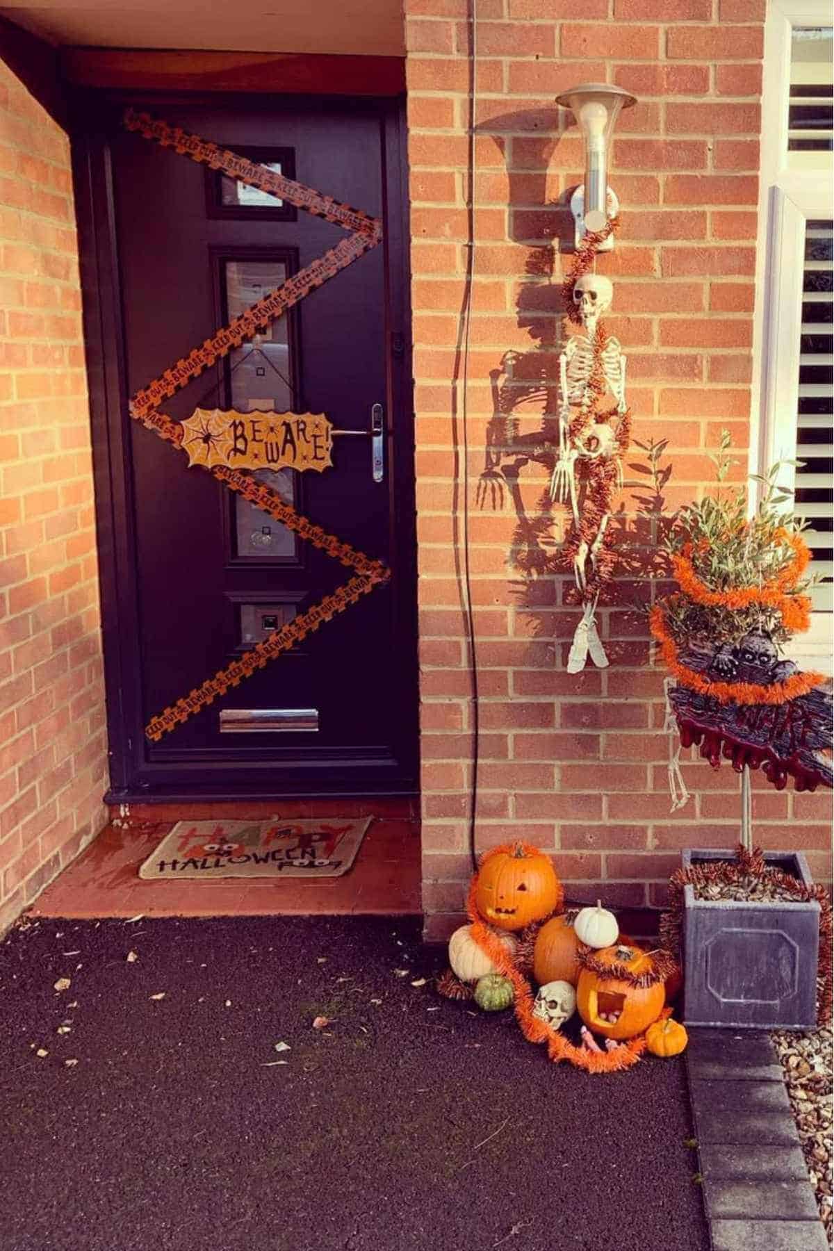 Front door decorated for Halloween with orange garlands, a "Beware" sign, a hanging skeleton, and a display of carved pumpkins and skulls beside a potted plant.