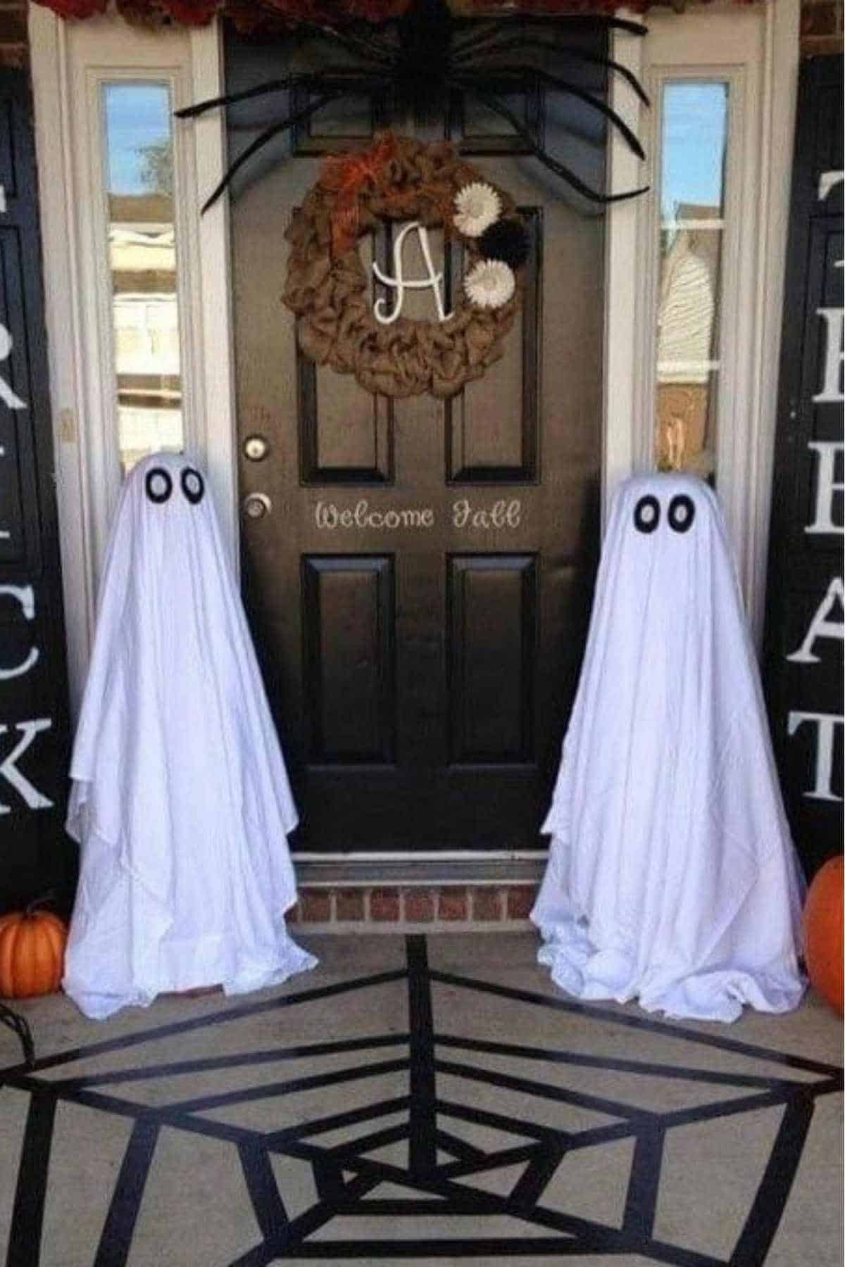 Two ghost decorations made from white sheets with black eyes stand by a black front door, which is decorated with a fall wreath and a spider. Pumpkins are on the porch.